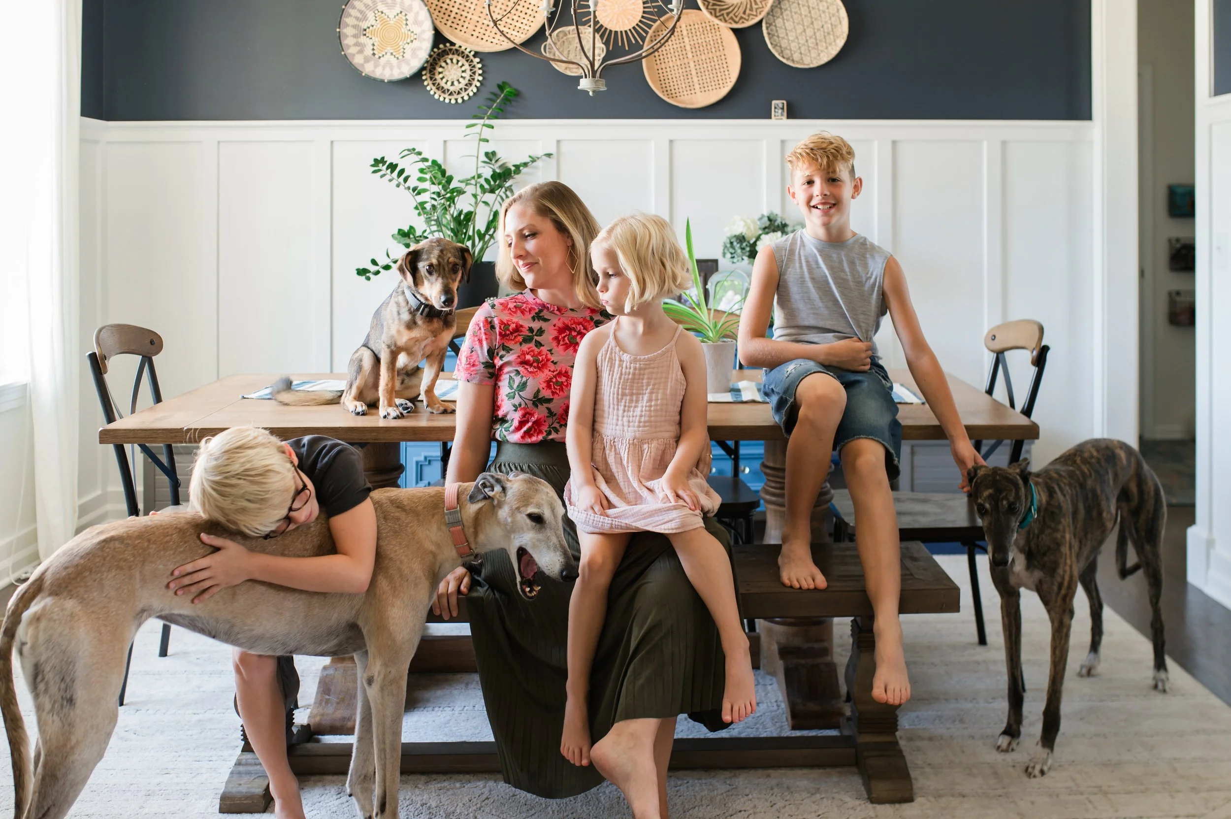 A family with children and dogs in a dining room, some children are sitting on a table while others and the dogs are around them, with wall decor and plants in the background.