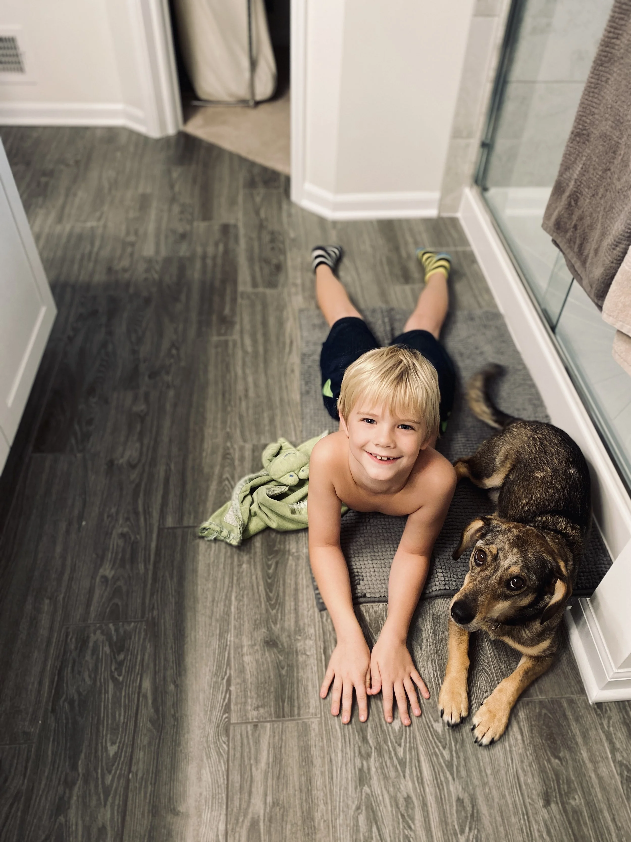 A young boy with blonde hair and a big smile lying on his stomach on the bathroom floor, with a dog next to him, and a towel hanging on the right side.