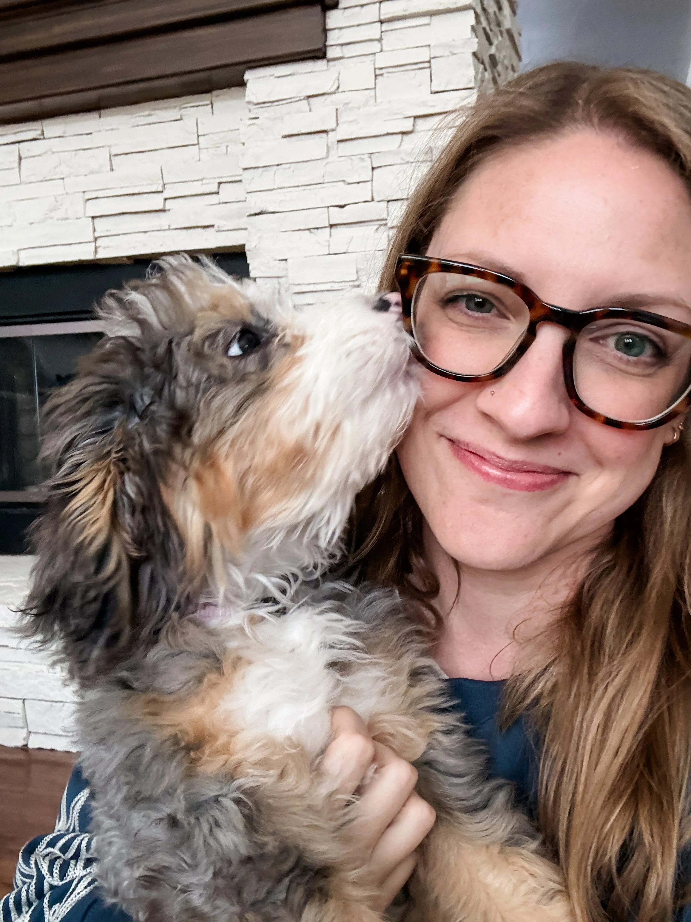 A woman with long, light brown hair, wearing glasses with a tortoiseshell pattern, is holding a fluffy dog with curly, multicolored fur. The woman is smiling gently and the dog is licking her face. The background features a wall with white stone brick and wood accents.
