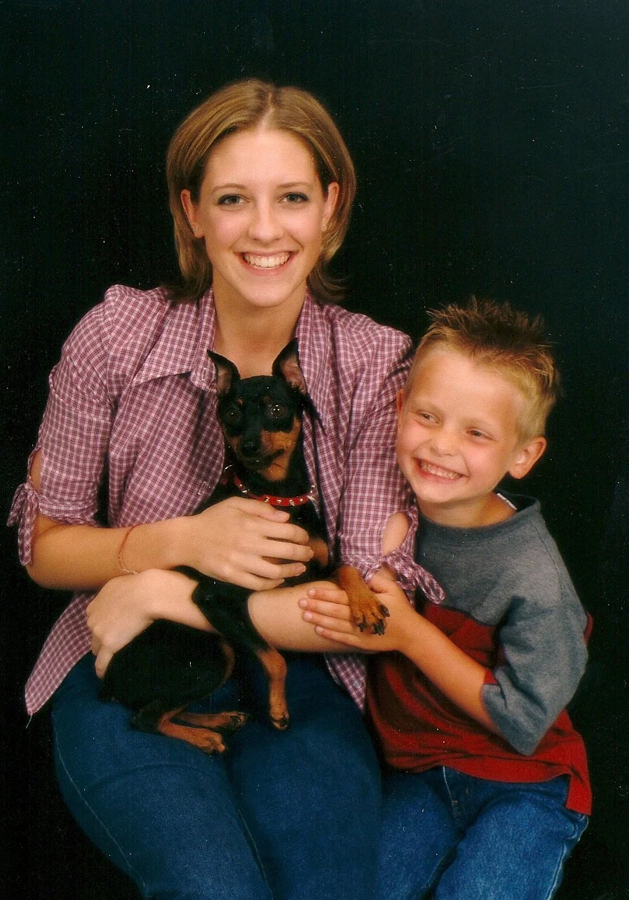 A woman and a young boy smiling and holding a small black and tan dog against a black background.