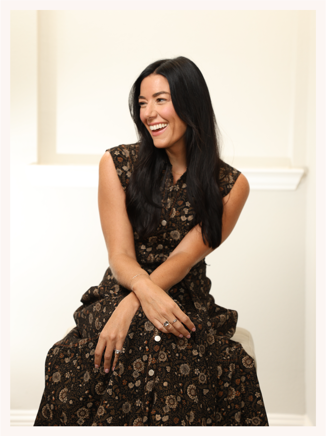 Woman with long black hair wearing a floral patterned dress, sitting and smiling indoors.