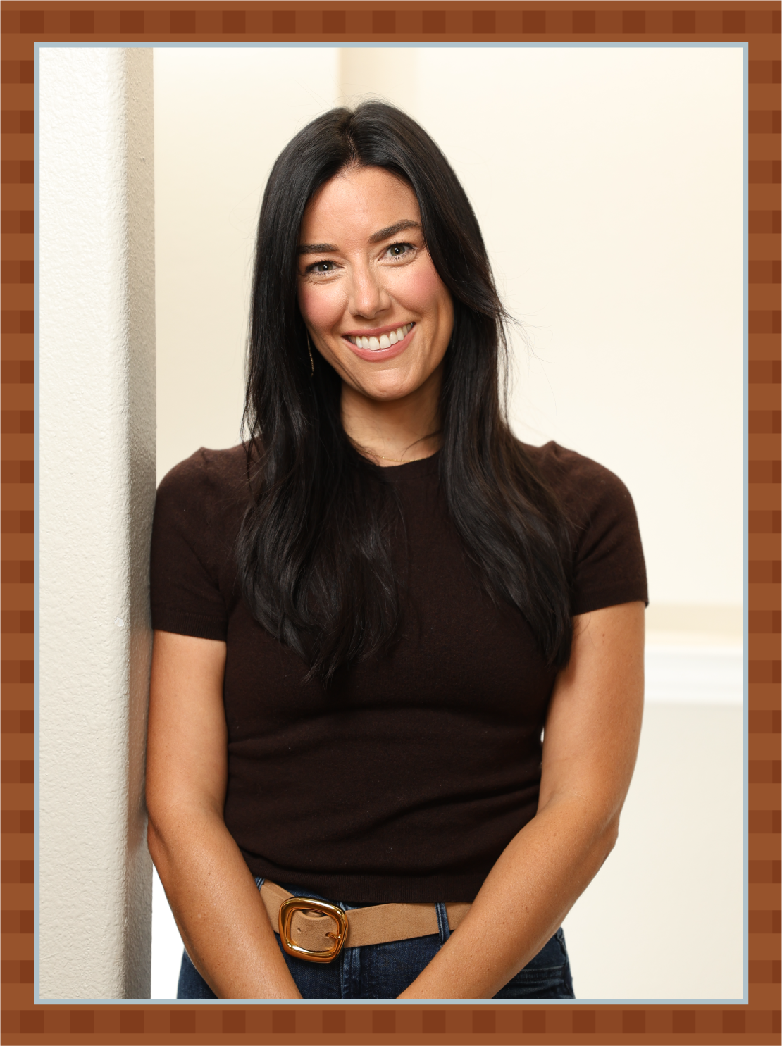 A woman with long dark hair wearing a brown top and a beige belt, smiling and leaning against a wall.