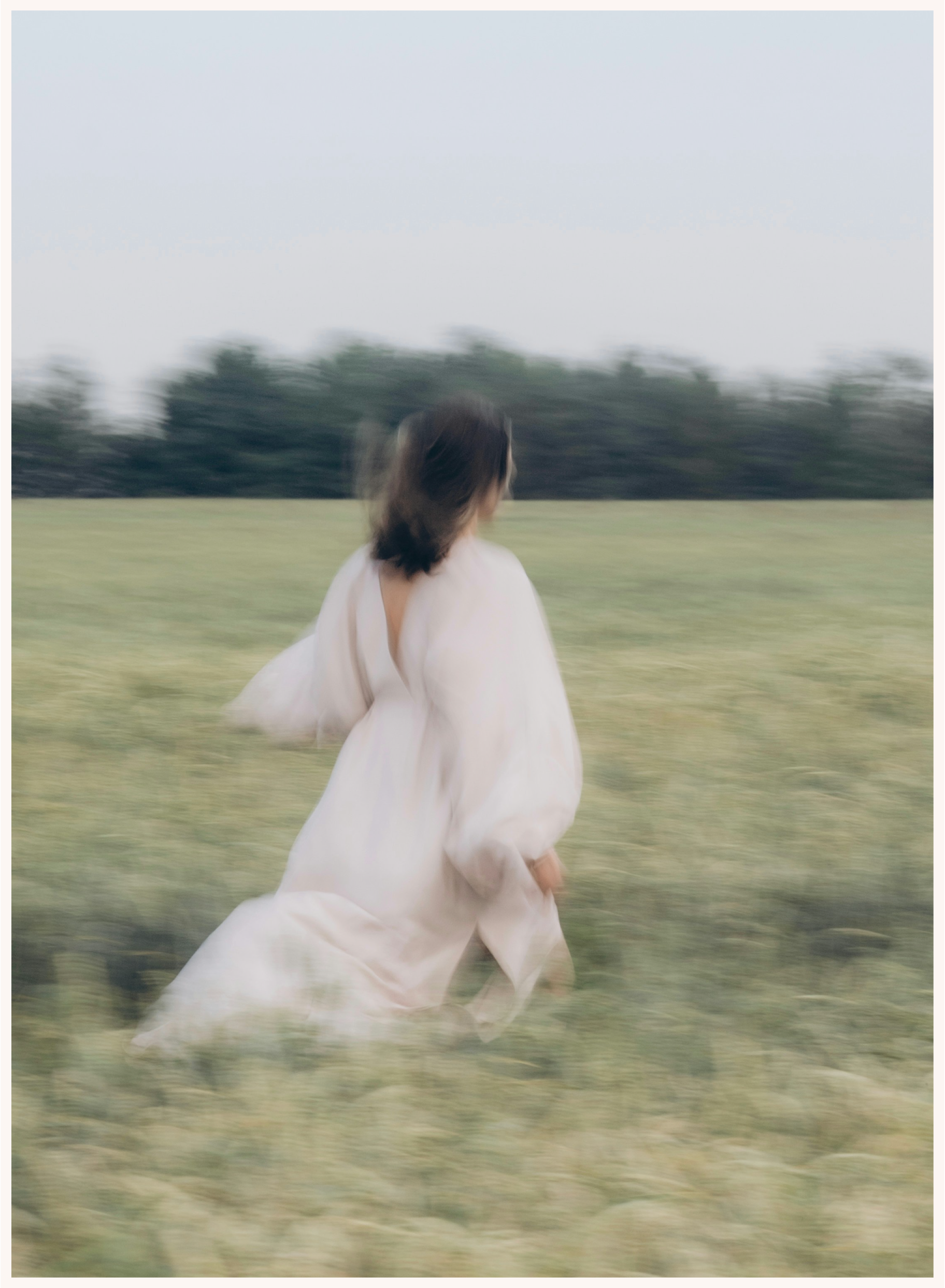 A person in a white dress running across a grassy field with trees in the background, captured with a motion blur effect.