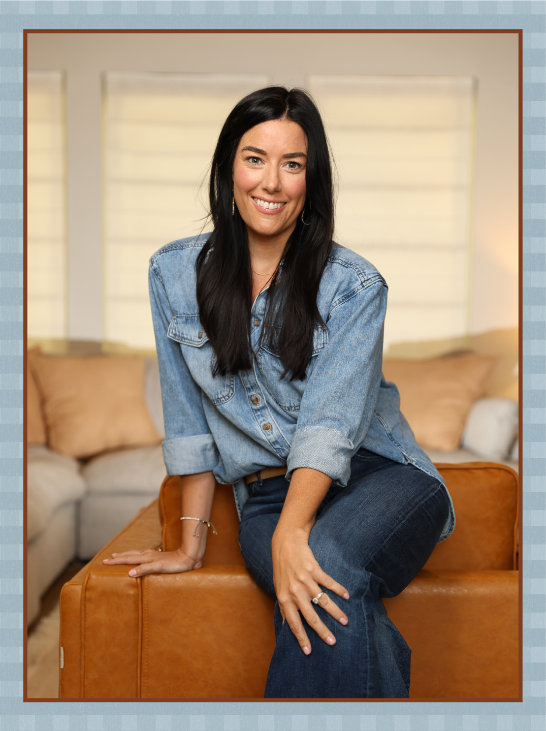 A woman with long dark hair wearing a denim jacket and jeans, smiling while sitting on an orange ottoman in a cozy living room with beige sofas and large windows.