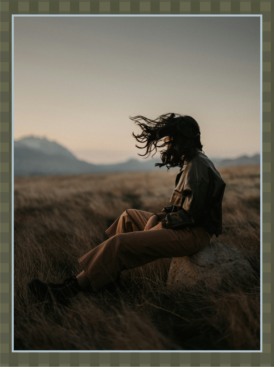 A woman with curly hair sitting on a rock in a grassy field at sunset or sunrise, with mountains in the background.