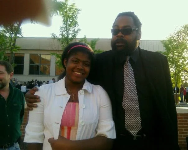 A woman with a pink headband and a man with sunglasses and a black suit, standing outdoors in front of a school building with other people in the background.