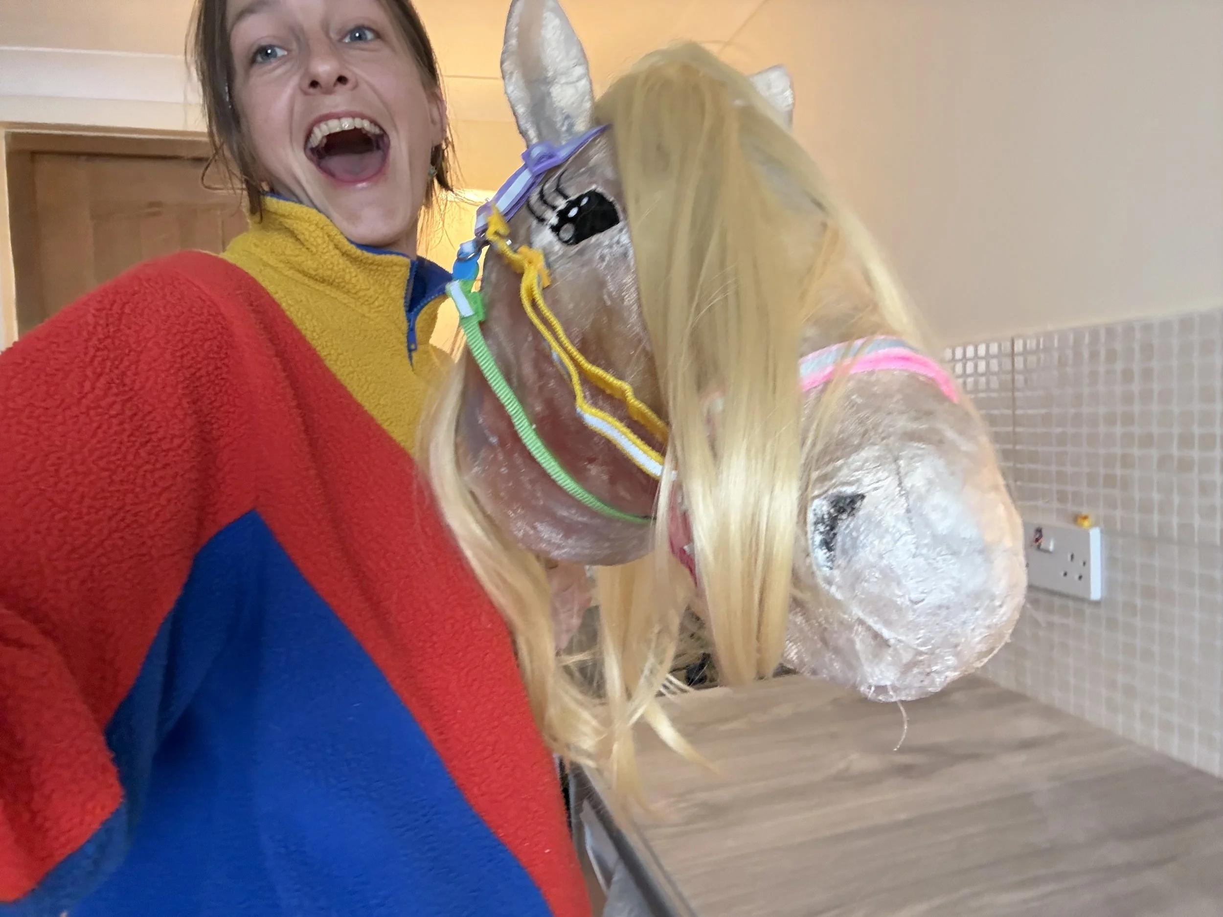 A stunningly gorgeous woman has a happy and excited expressions, eyes sparkling and mouth open smiling. she holds a life size horse head made of plaster bandage that she crafted herself. he looks very fabulous and realistic. he is painted gold.