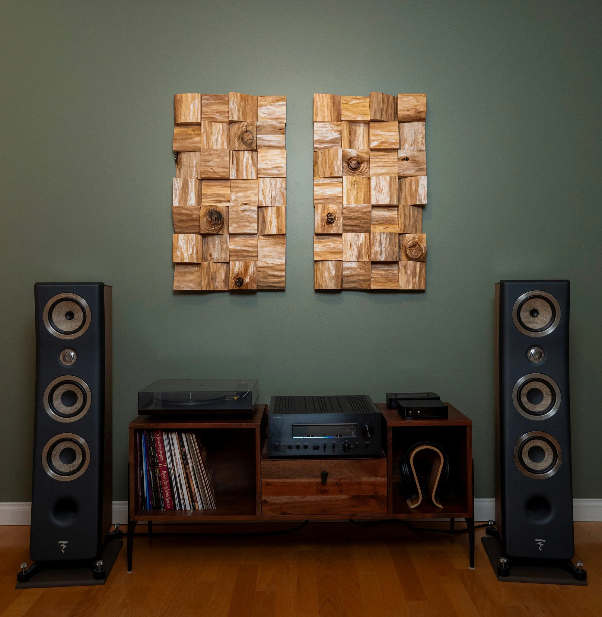 A home audio setup with two large floor-standing speakers, a wooden media console holding vinyl records, an audio receiver, and a turntable, all against a green wall with wooden flooring.
