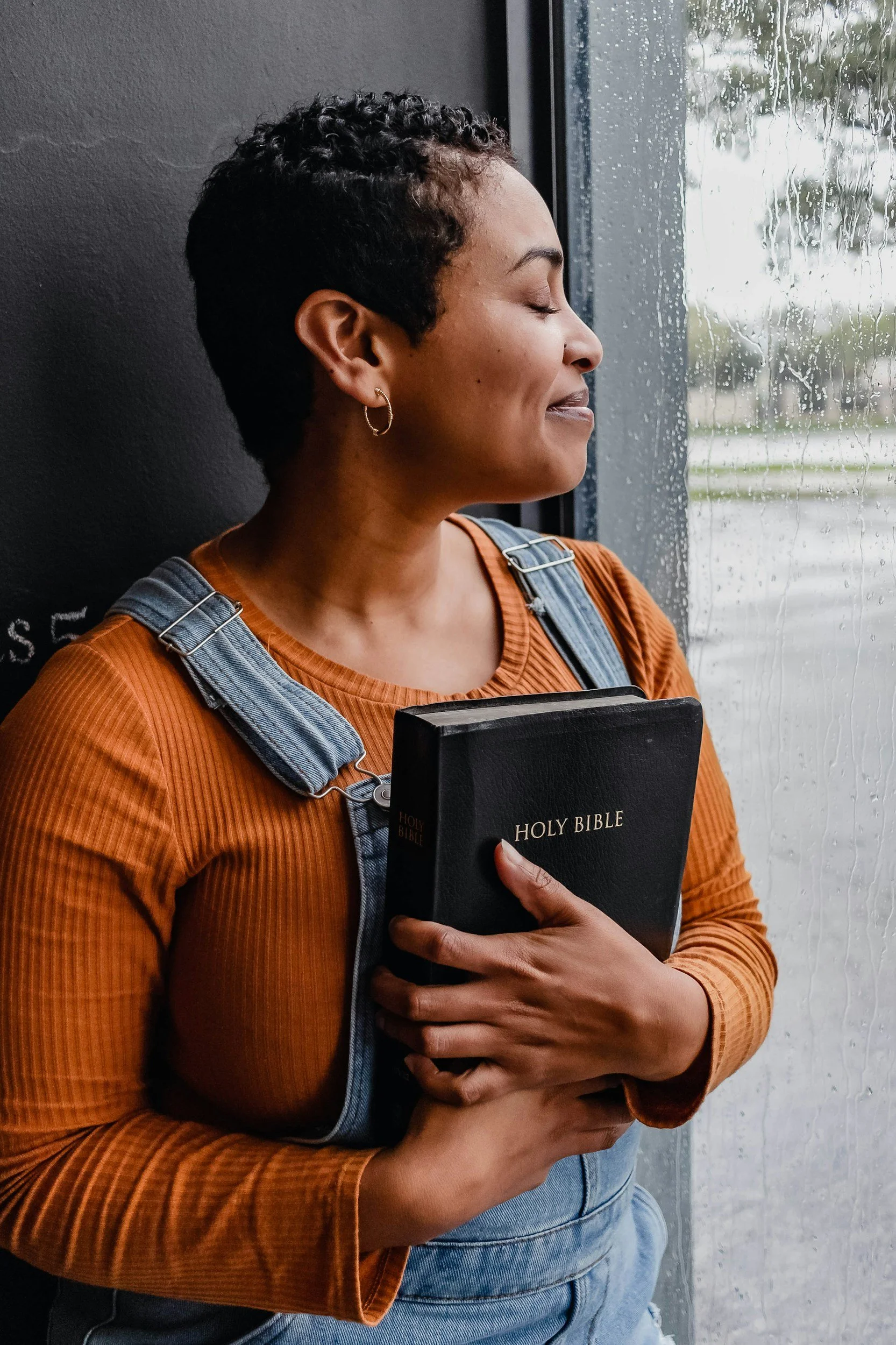 A woman with short, curly black hair, wearing earrings, an orange ribbed long sleeve shirt, and denim overalls, holding a black Holy Bible close to her chest, standing by a window with rain on the glass.