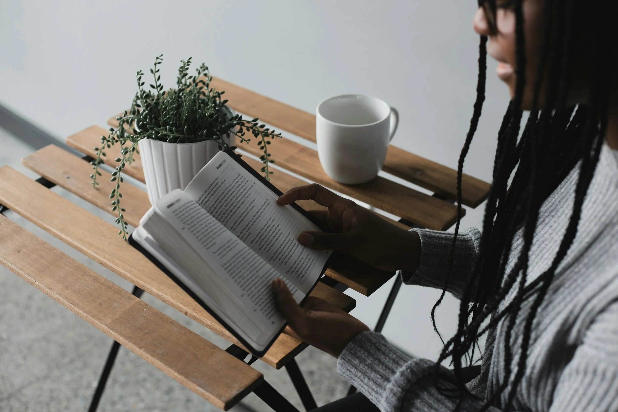 A person with long braids is sitting at a wooden table, reading a book and holding it with both hands, next to a white mug and a potted plant.