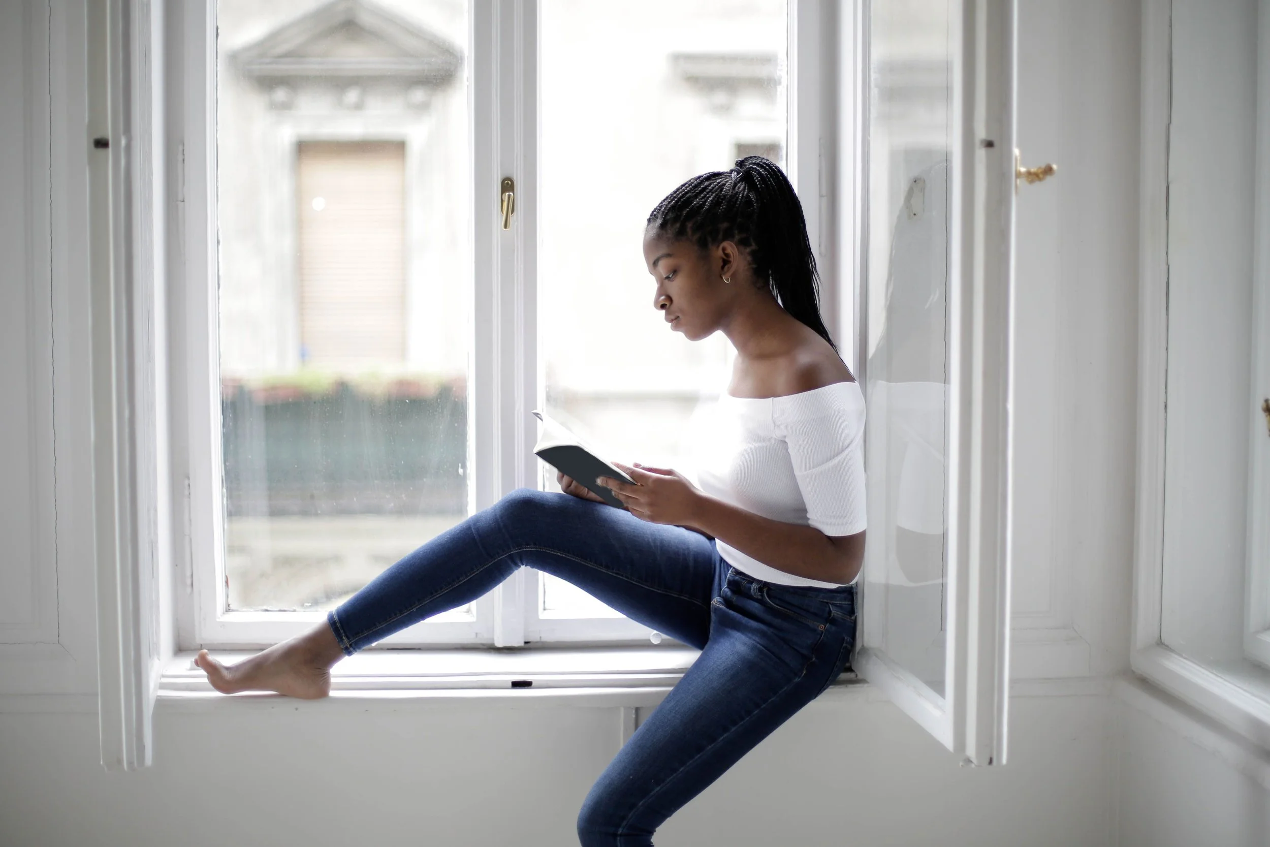 A young woman with braided hair sitting on a wide window sill, reading a book, with her legs stretched out and crossed at the ankles. She is wearing a white off-the-shoulder top and blue jeans, inside a bright room.