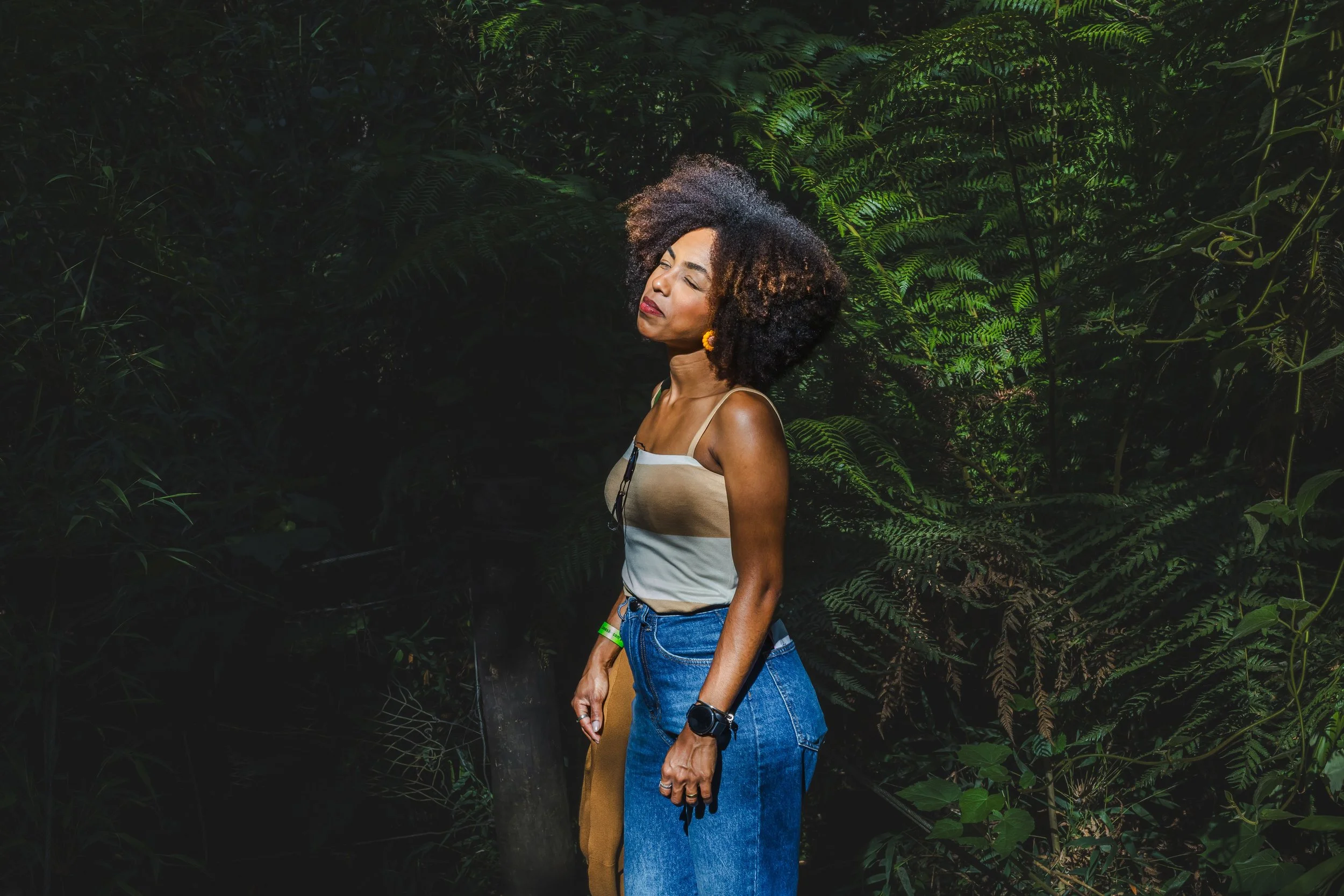 A woman with curly hair closed her eyes as she stands in front of lush green foliage in a jungle or forest setting.