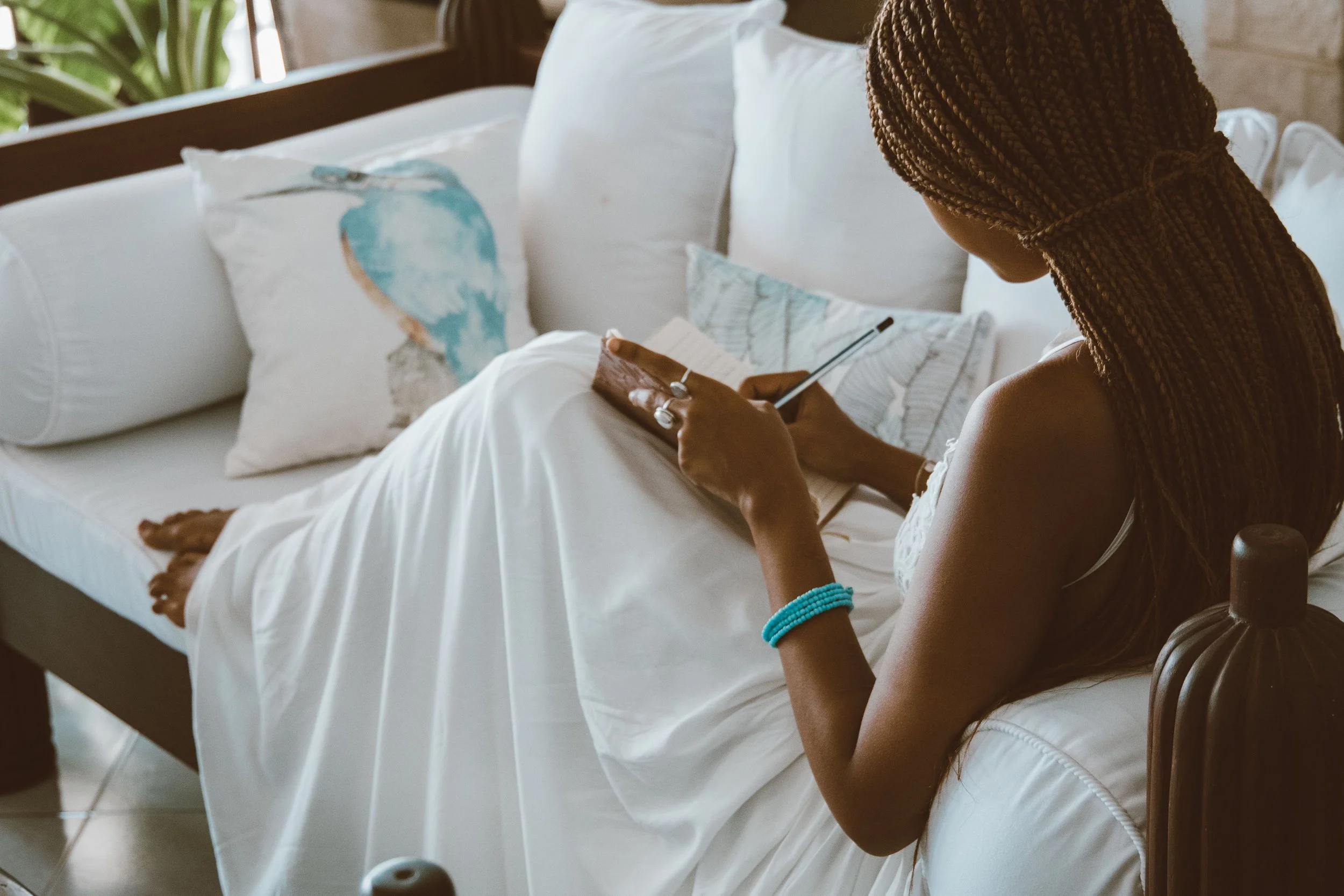 A woman with braided hair and turquoise bracelets sitting beside a bed, looking at her phone, with pillows and a painting of a bird in the background.