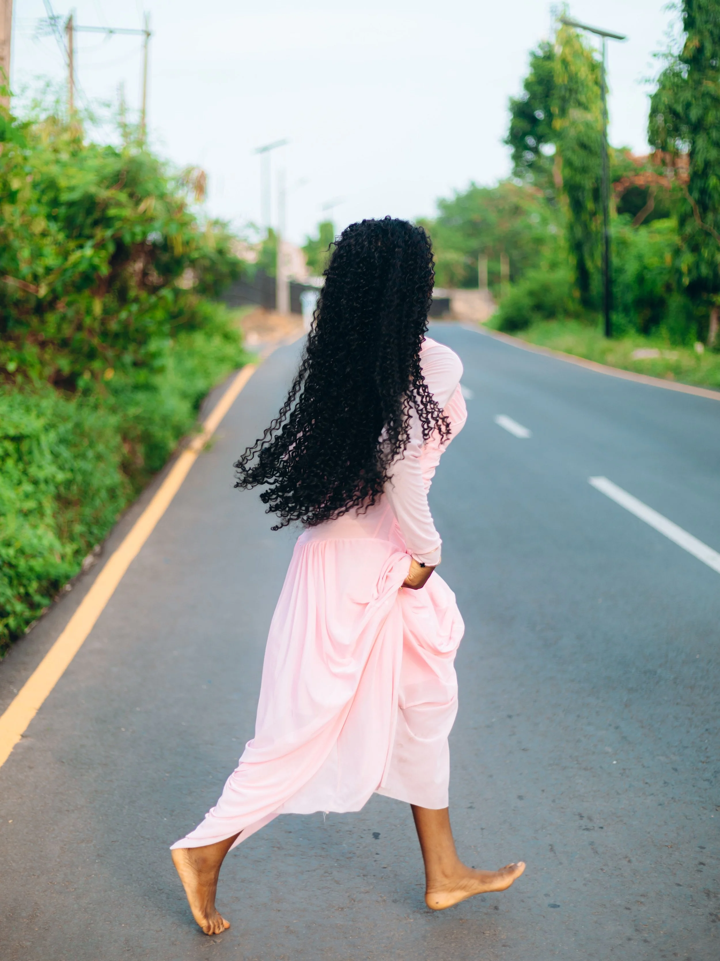 A woman with long, curly black hair in a pink dress standing on a paved road surrounded by green trees and shrubbery.