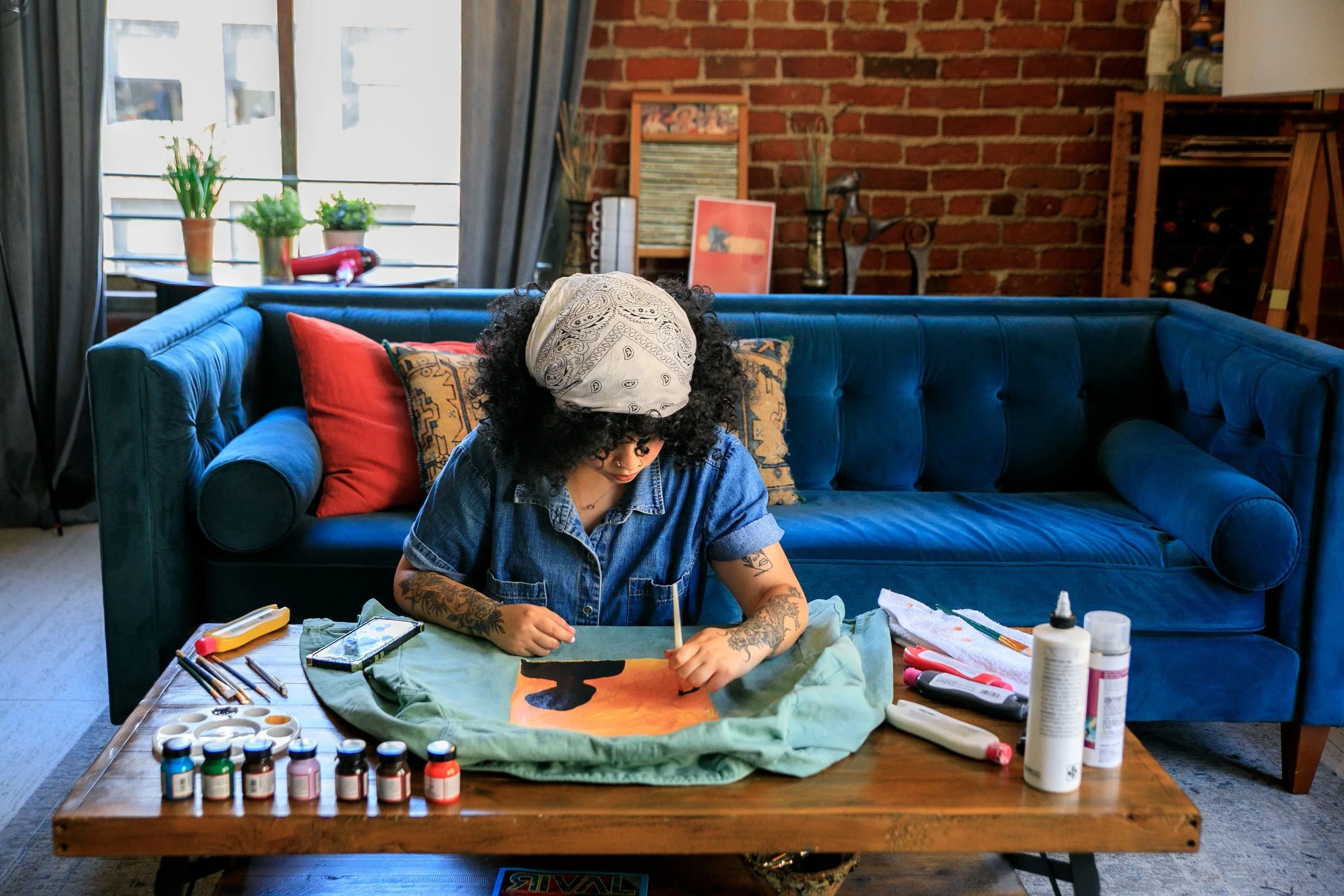 A woman with curly hair, tattoos, and a bandana working on a colorful abstract painting at a cluttered table in a cozy living room with exposed brick walls, a blue couch, and various art supplies.
