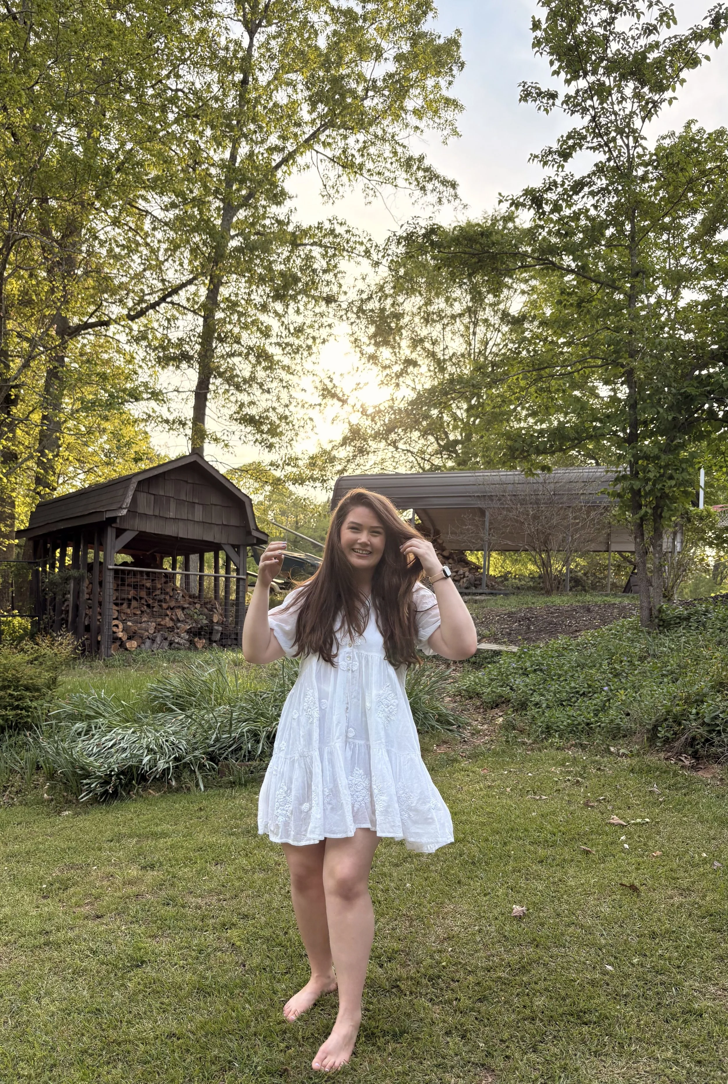 A young woman in a white dress standing barefoot on grass in a backyard, smiling, with trees, a wooden shed, and a covered patio in the background during late afternoon or early evening.