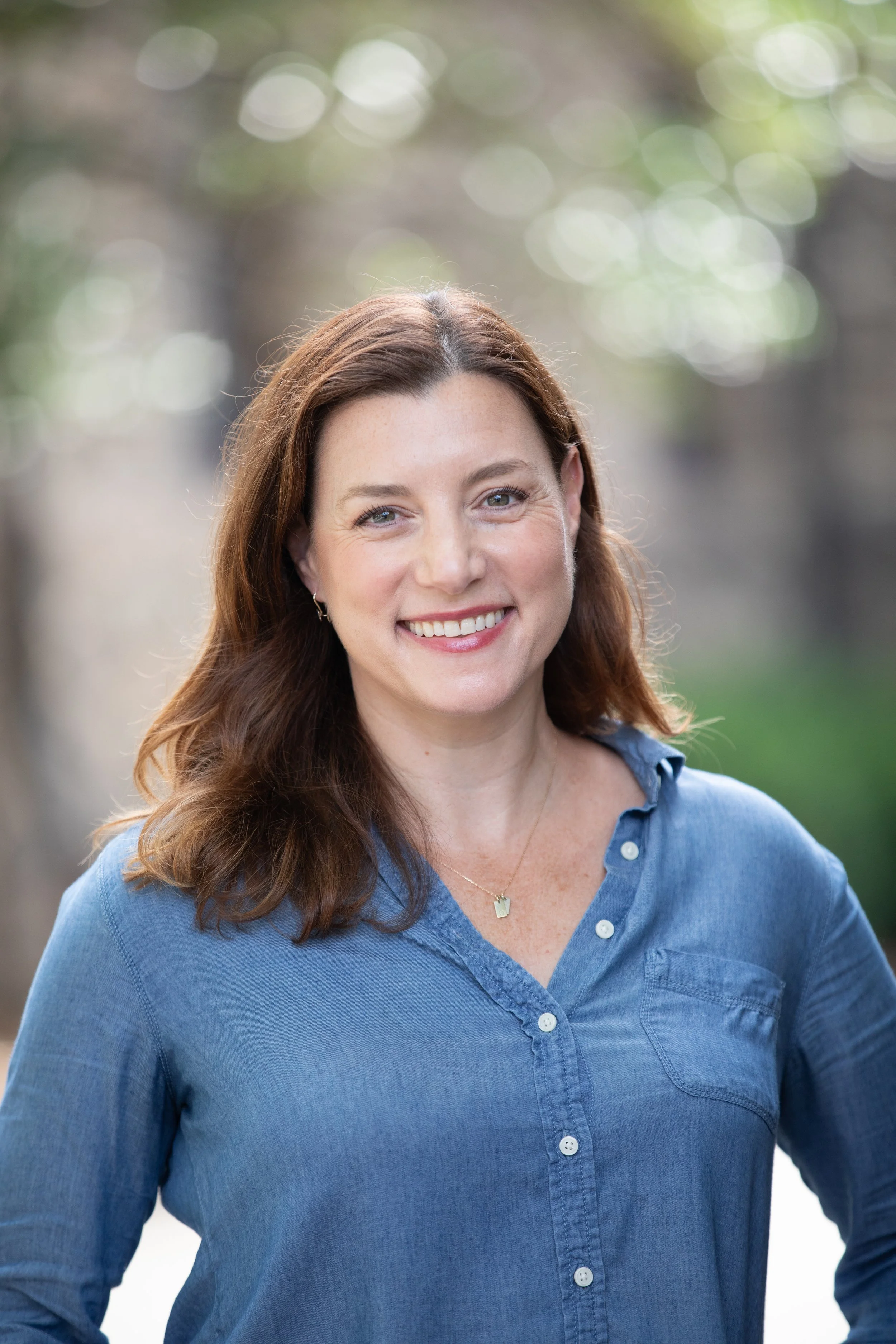 Close-up of a smiling woman with long, wavy brown hair, wearing a light blue button-up shirt and a gold necklace, outdoors with blurred greenery in the background.