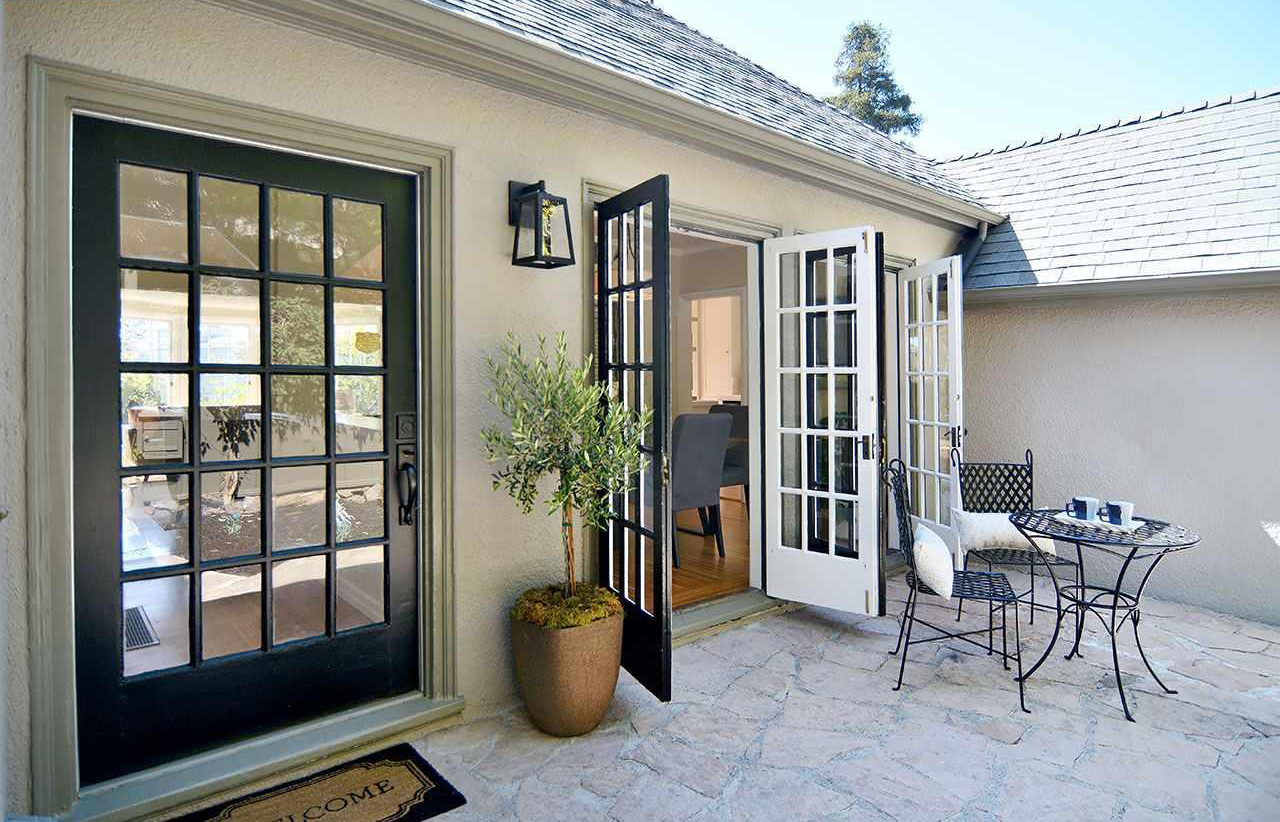 A small outdoor patio area with a metal table and two chairs, a potted plant, and a set of open French doors leading into a house.