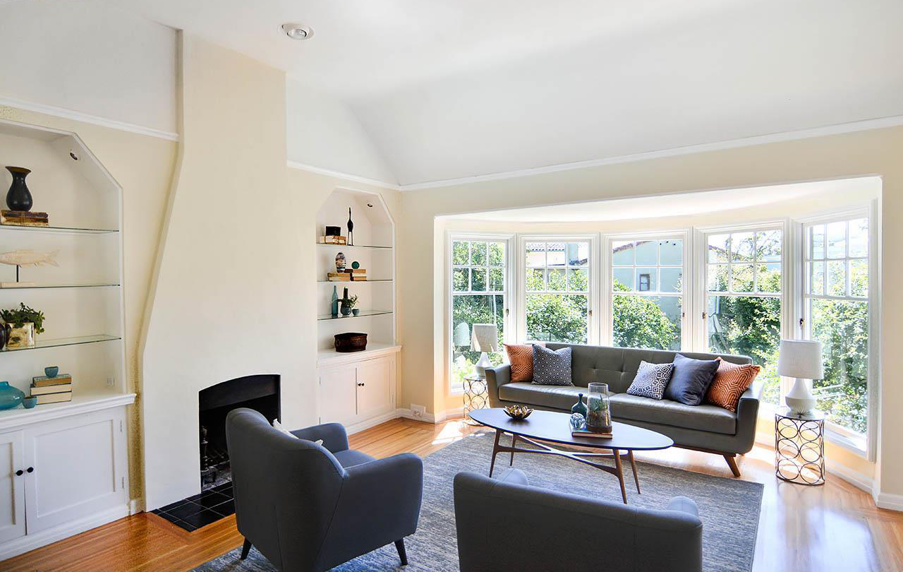 Bright living room with large bay window, gray sofa, two gray armchairs, wooden coffee table, and built-in shelves with decorative items.