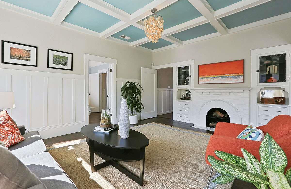 A living room with white paneled walls, a white brick fireplace, colorful artwork, a gray couch with patterned pillows, an orange armchair with a leafy throw pillow, a black coffee table with vases, a large plant in a white pot, and a coffered ceiling with a chandelier.
