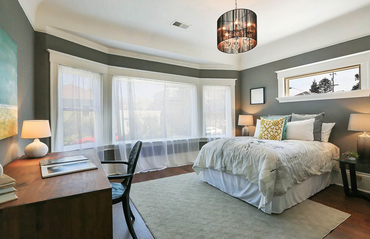 Bright bedroom with gray walls, a large window with sheer curtains, a bed with white and decorative pillows, side tables with lamps, a wooden desk with a lamp and books, and a chandelier ceiling light.