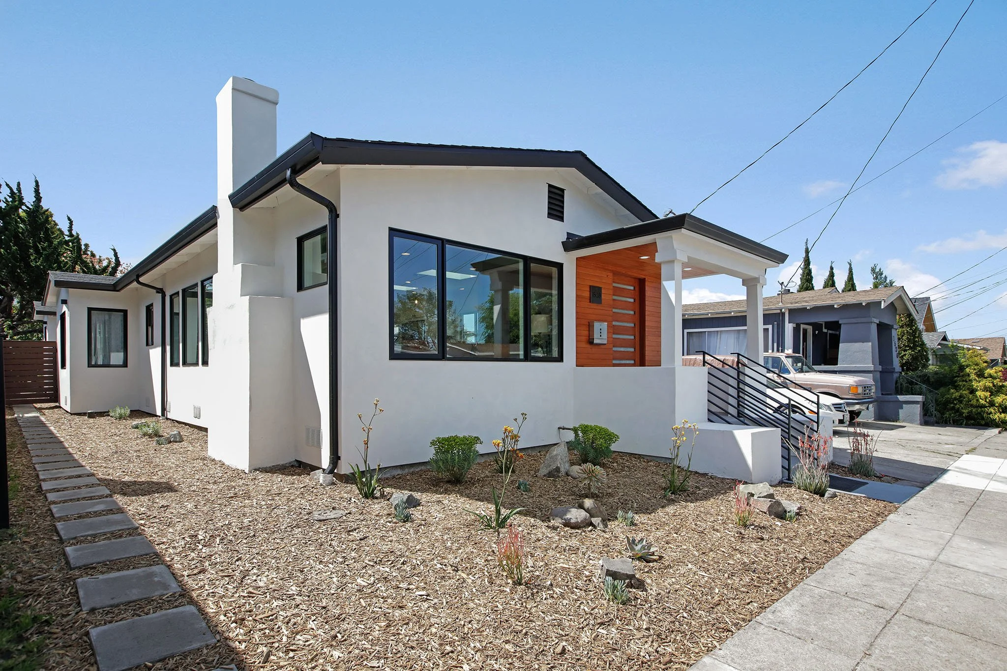 Front view of a modern white house with black window frames and a small porch, a driveway with parked cars, and a landscaped yard with desert plants and rocks.