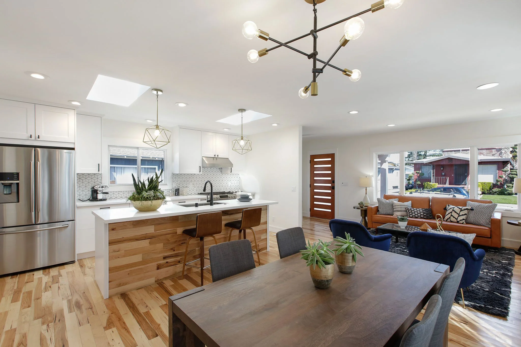 Open-concept kitchen and living room with modern lighting, white cabinetry, stainless steel refrigerator, dining table, and large windows with outdoor views.