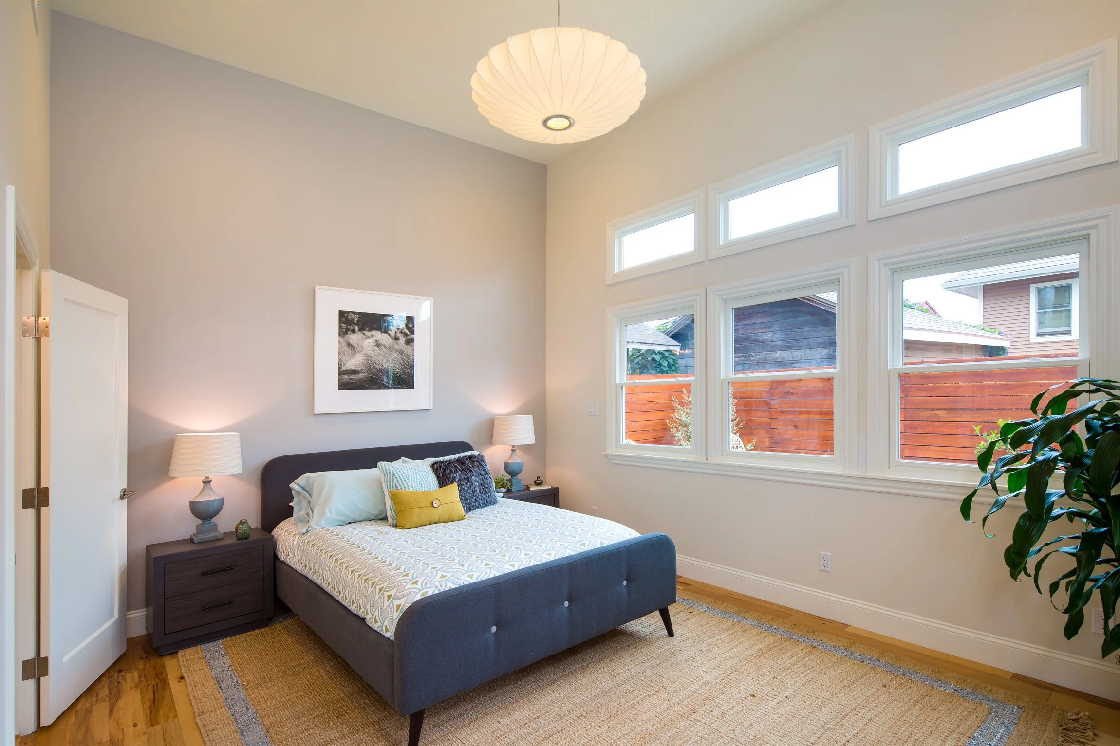 Bedroom with a bed, nightstand, lamps, and large windows showing an outdoor fence and neighboring house.