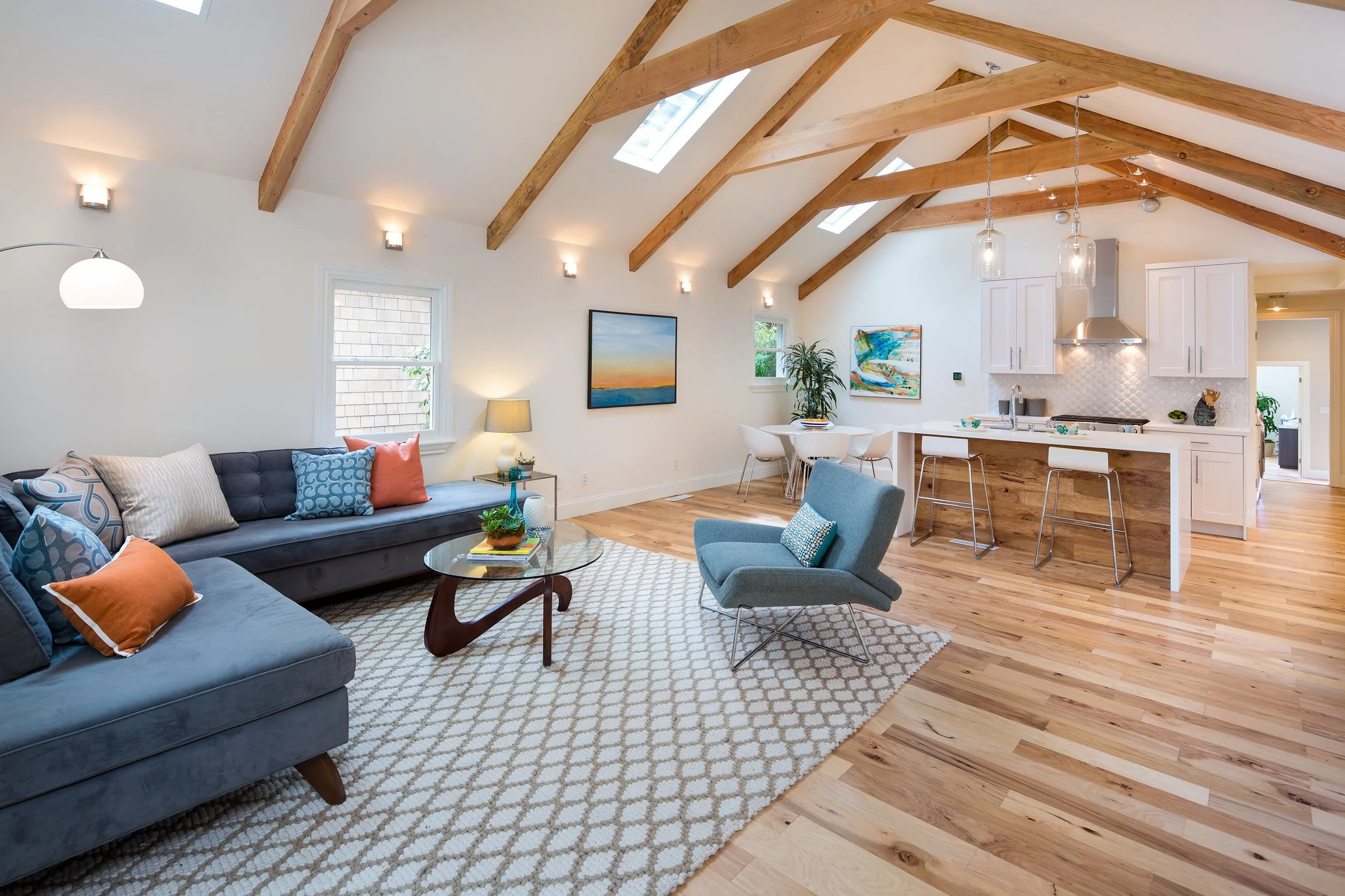 A spacious living room with vaulted ceiling and exposed wooden beams, featuring large windows, a blue sofa with colorful cushions, a round glass coffee table, a patterned rug, a light blue armchair, and wall art, connected to the kitchen area with white cabinetry and pendant lighting.