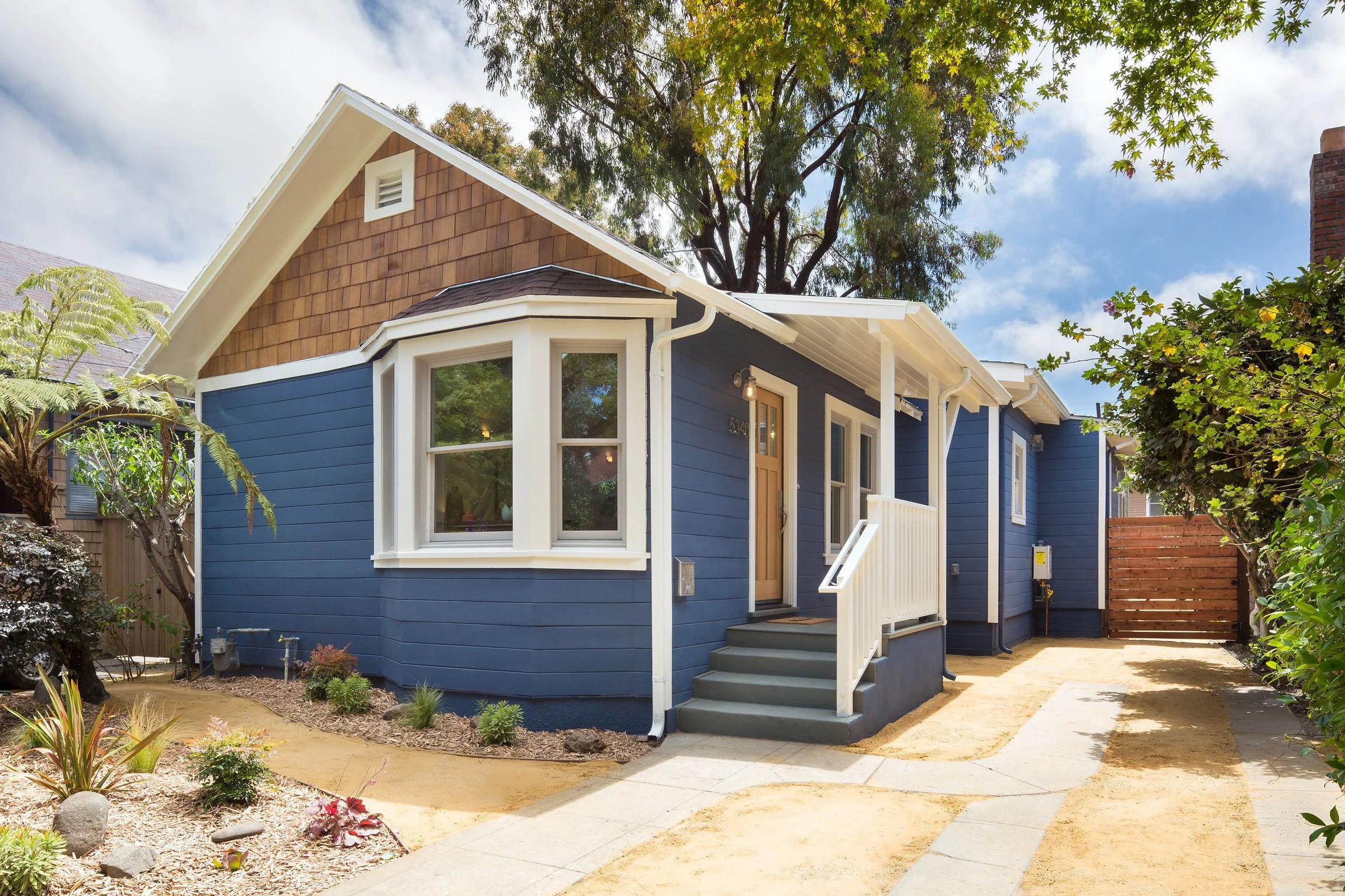 Front view of a blue house with a brown-shingled upper wall, white trim, and front porch steps, surrounded by trees and plants, under a partly cloudy sky.