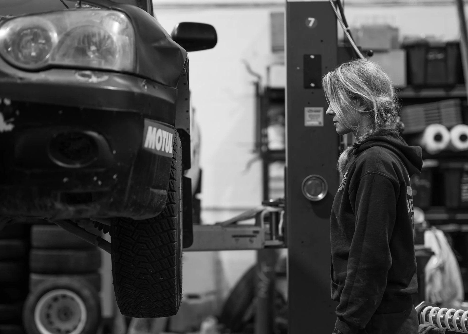 A woman with glasses and long hair in a ponytail working on a car in a garage or workshop. The car is lifted, and she appears to be inspecting or working on the wheel area.