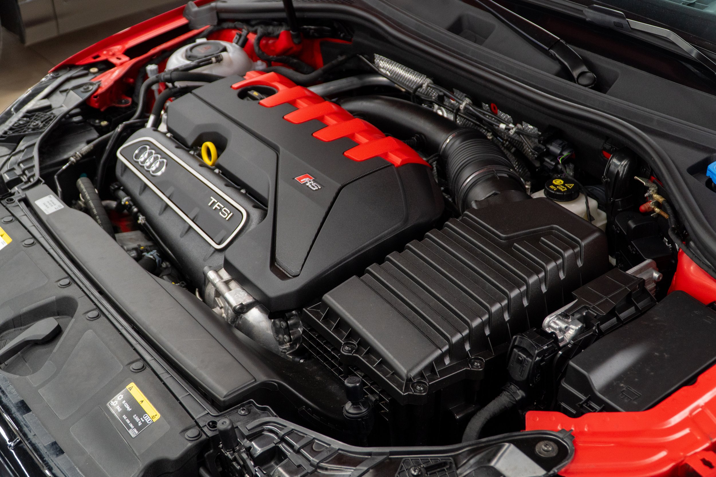 A car engine bay with a V6 TFSI engine, featuring black and red components, hoses, and electrical wiring.