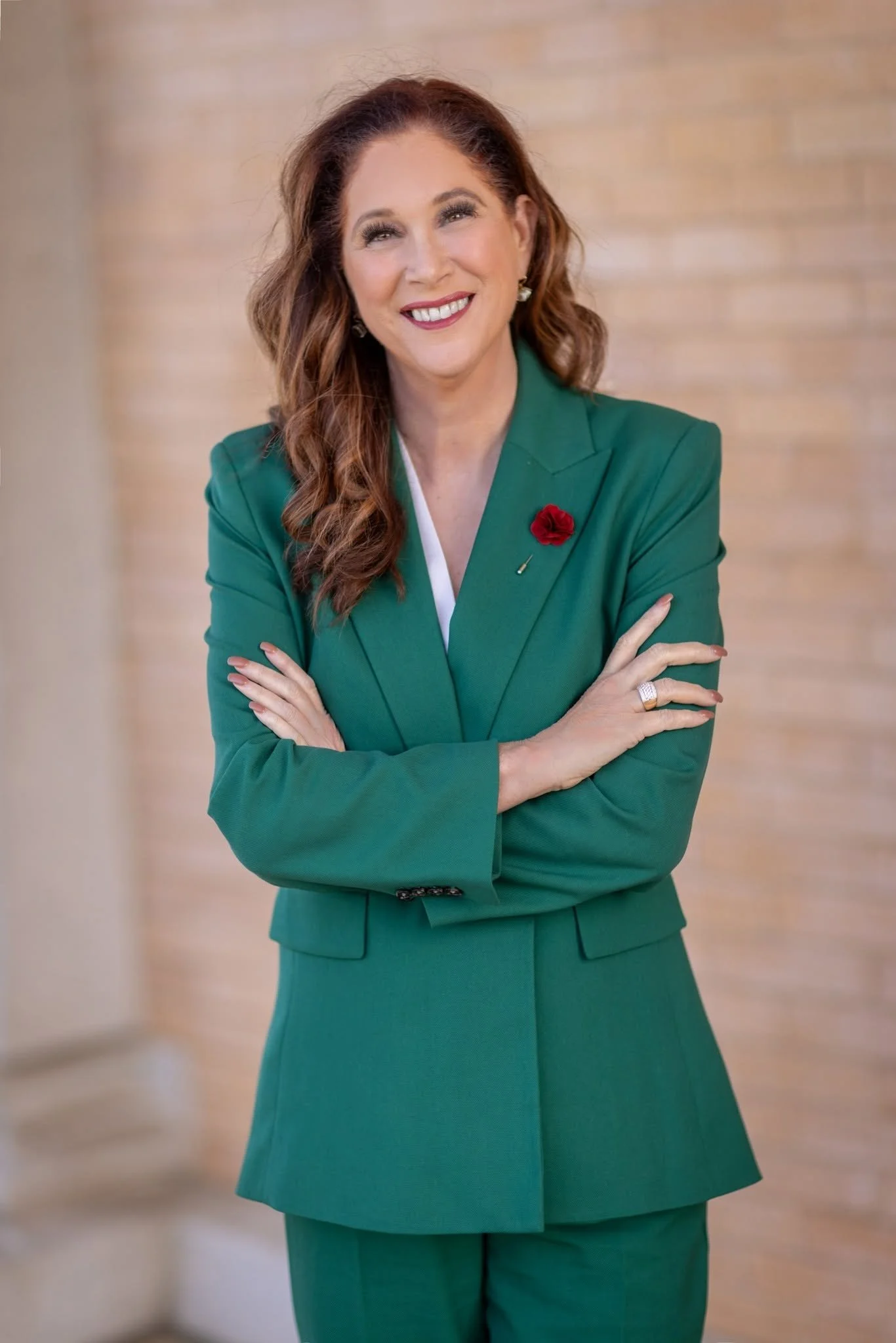 A woman with long, wavy reddish-brown hair and fair skin, smiling, wearing a tailored green blazer with a red flower pin and matching green skirt, standing arms crossed against a beige-colored wall.