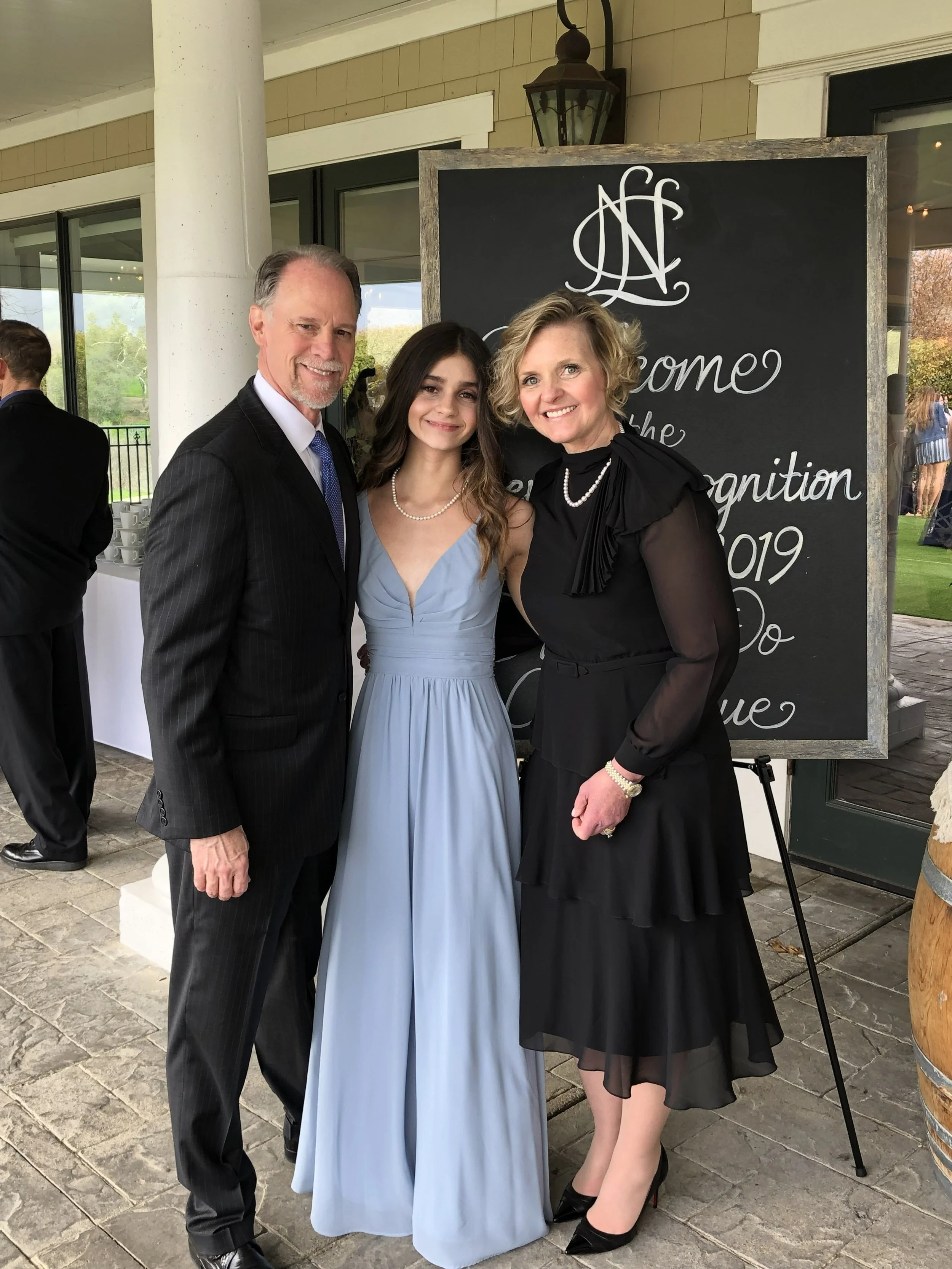 Three people dressed in formal attire standing and smiling outside during daytime. There is a man in a dark suit, a woman in a light blue gown, and a woman in a black dress with sheer sleeves. Behind them, there is a large black chalkboard with writing on it, and a window showing a green outdoor area.