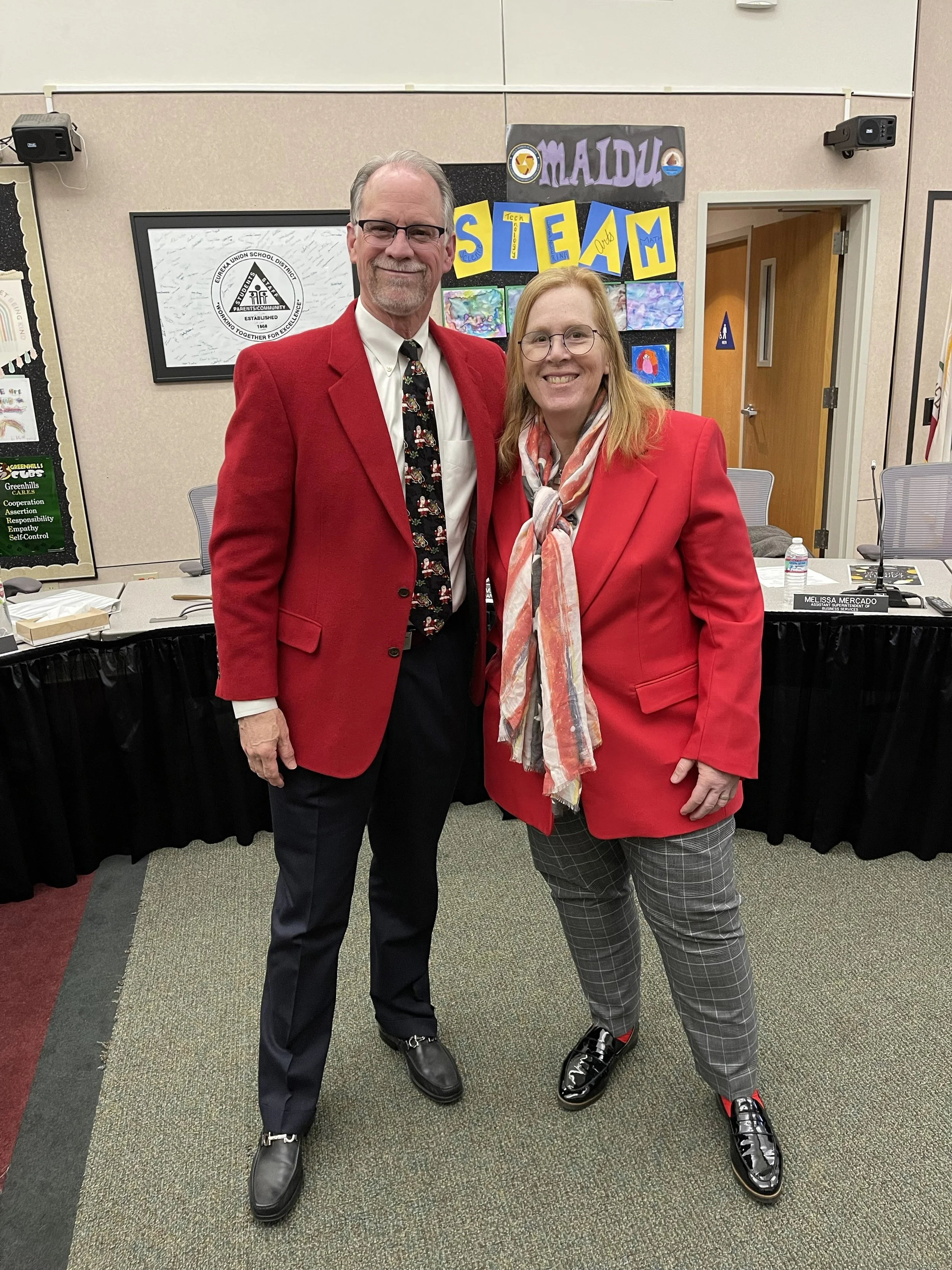 Two people standing together in a room with posters on the wall and tables. One person is a man wearing glasses, a red blazer, a white shirt, an animated tie, black pants, and black shoes. The other person is a woman with glasses, long hair, and a red blazer over a patterned shirt and plaid pants, wearing shiny black shoes.