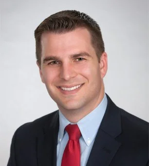 A professional headshot of a man in a navy suit, white shirt, and red tie, smiling against a light gray background.