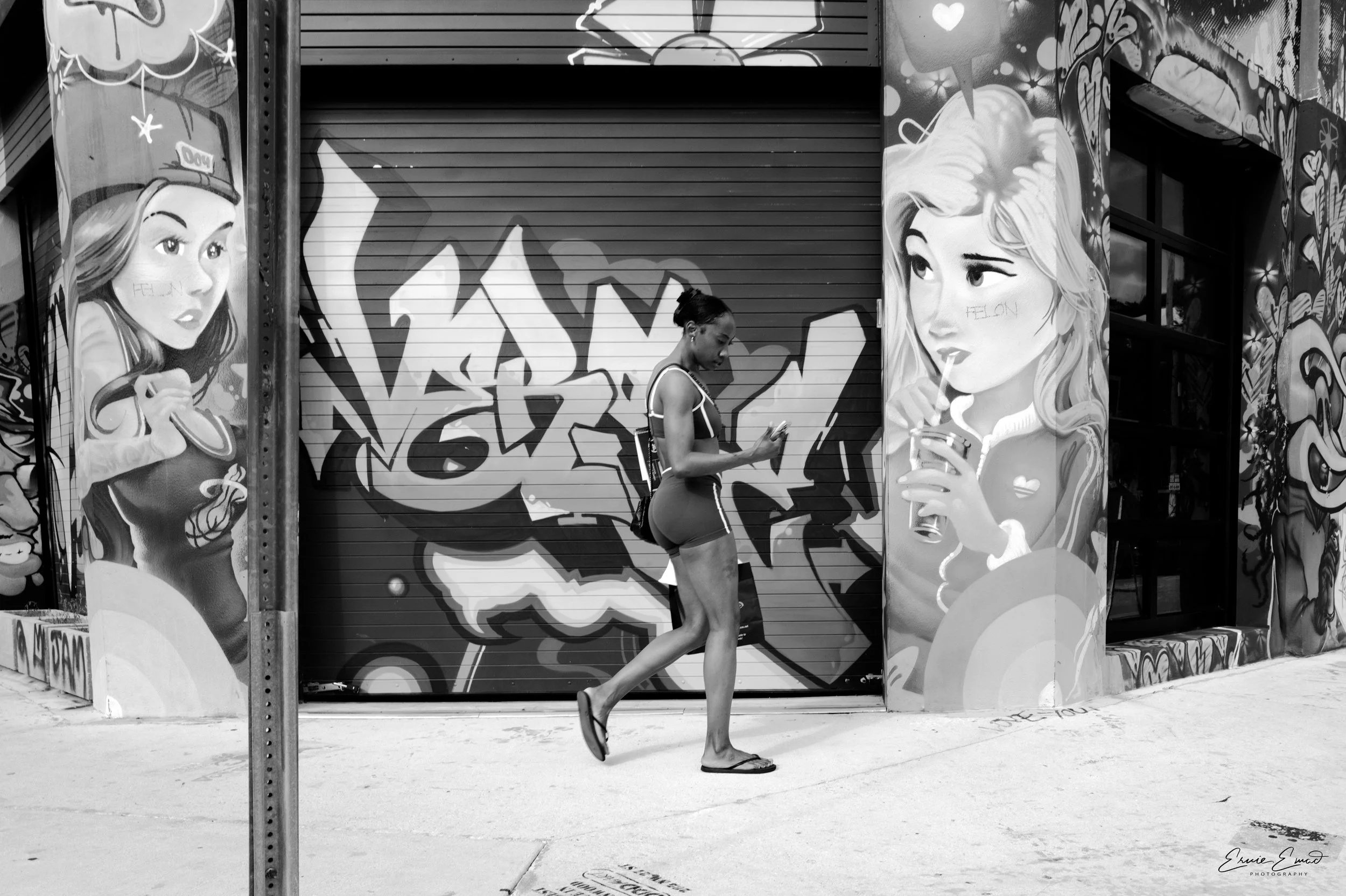 A young woman with a ponytail walking on the sidewalk while looking at her phone, passing by graffiti art on metal garage doors featuring animated women holding drinks.