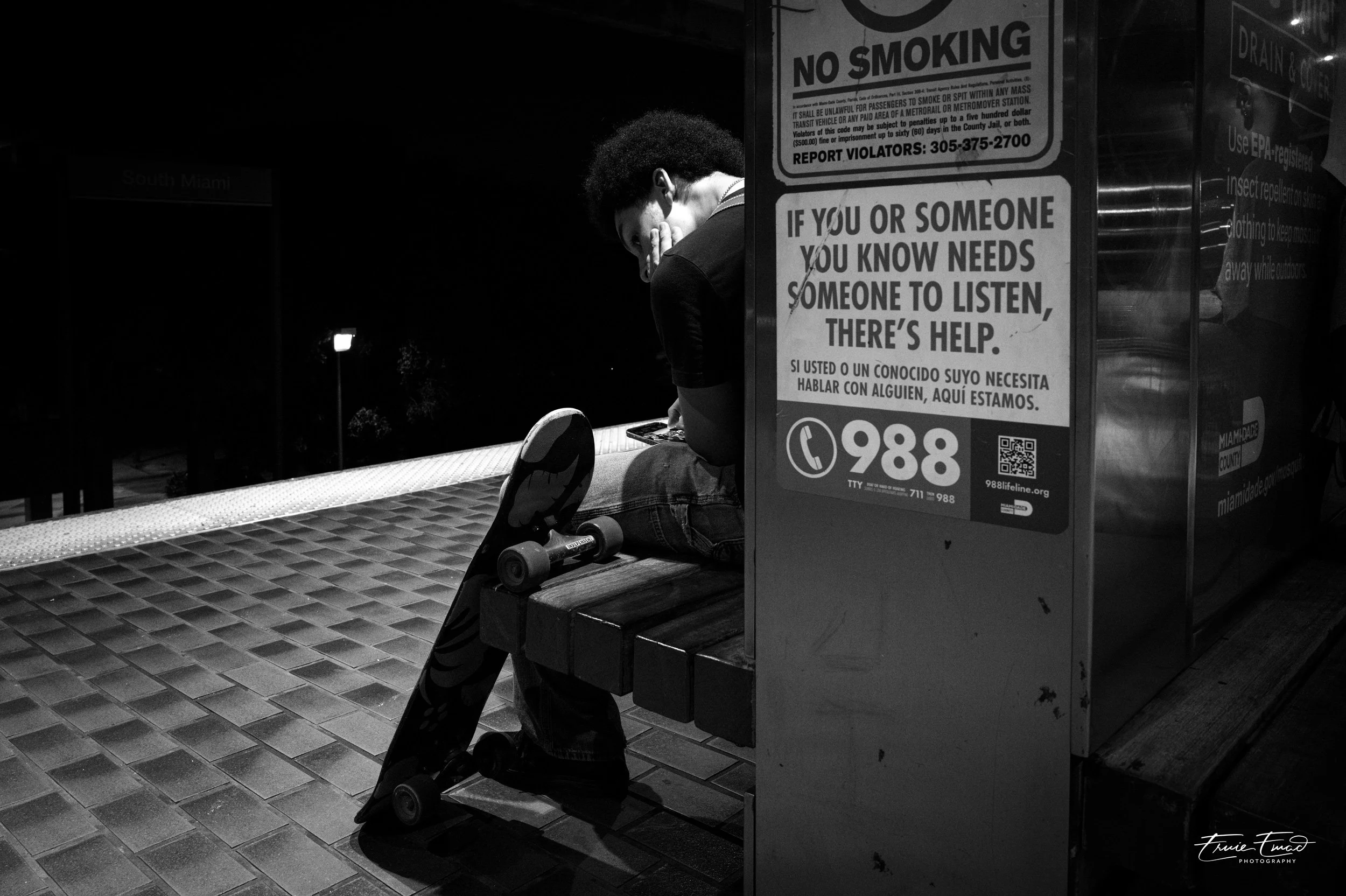 A person sitting on a bench at night, wearing sunglasses, with a skateboard resting against the bench. There are several signs nearby, including a 'No Smoking' sign and a message about helping others in need. The area appears to be a transit station,