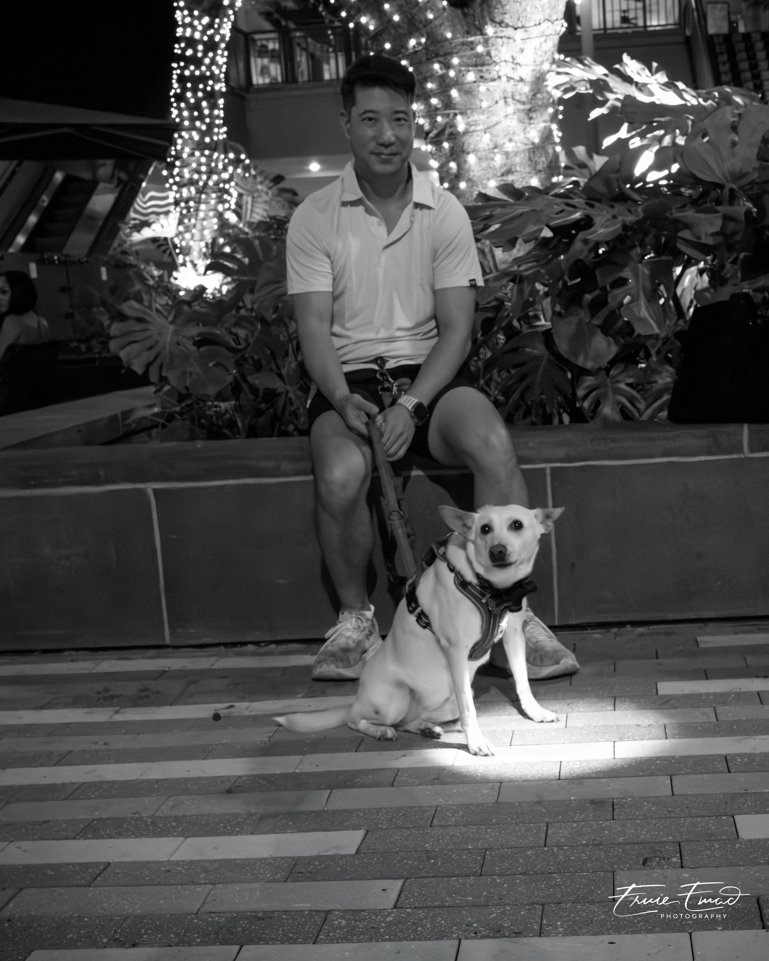 A man in a white shirt sitting on a bench in front of large leafy plants and illuminated trees at night, with a dog sitting in front of him. The image is in black and white.