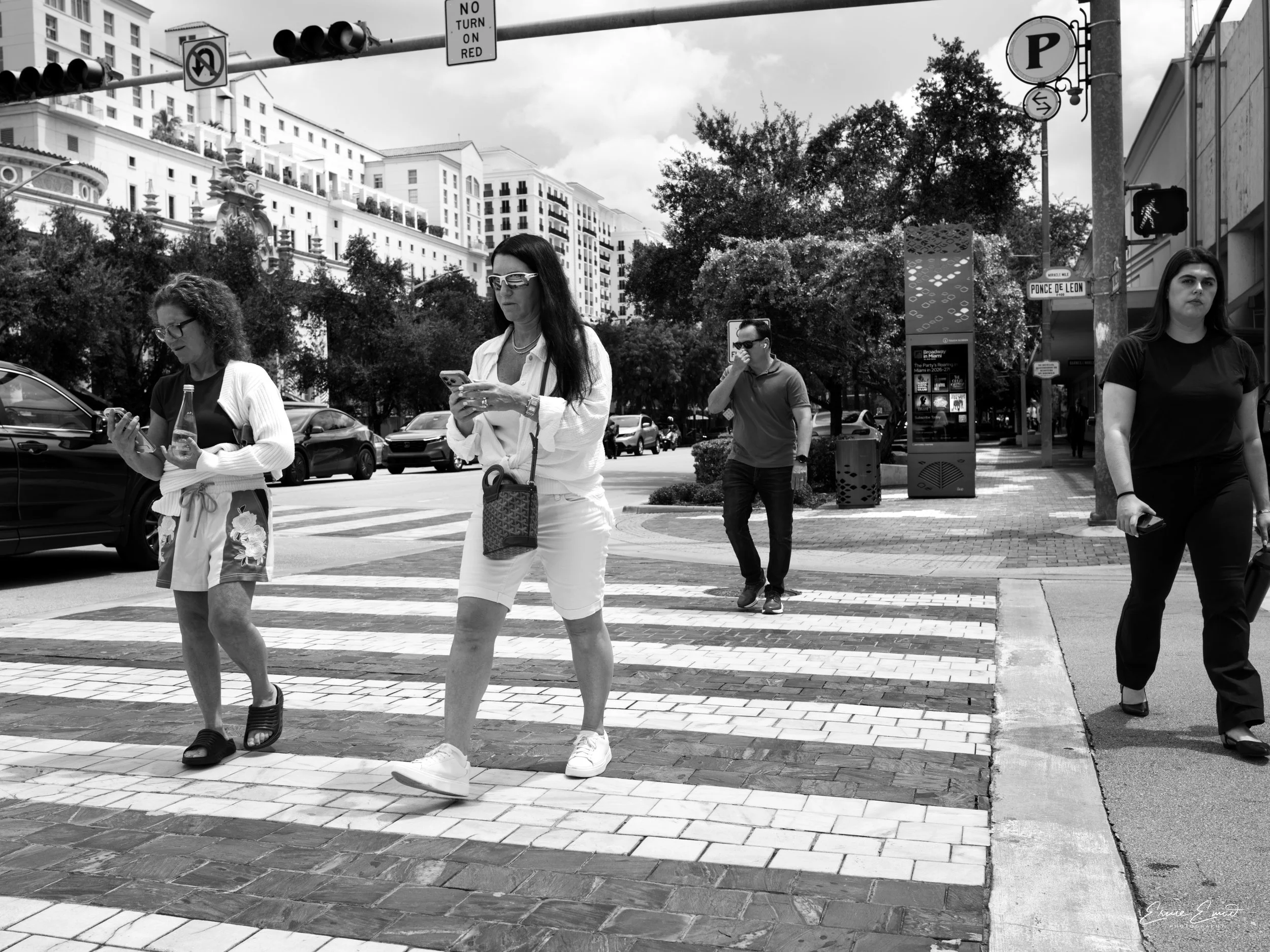 Black and white photo of four people crossing a city street at a crosswalk, with tall buildings and trees in the background. Two women in the foreground are looking at their phones, and a man and another woman are walking behind them on Ponce de Leon