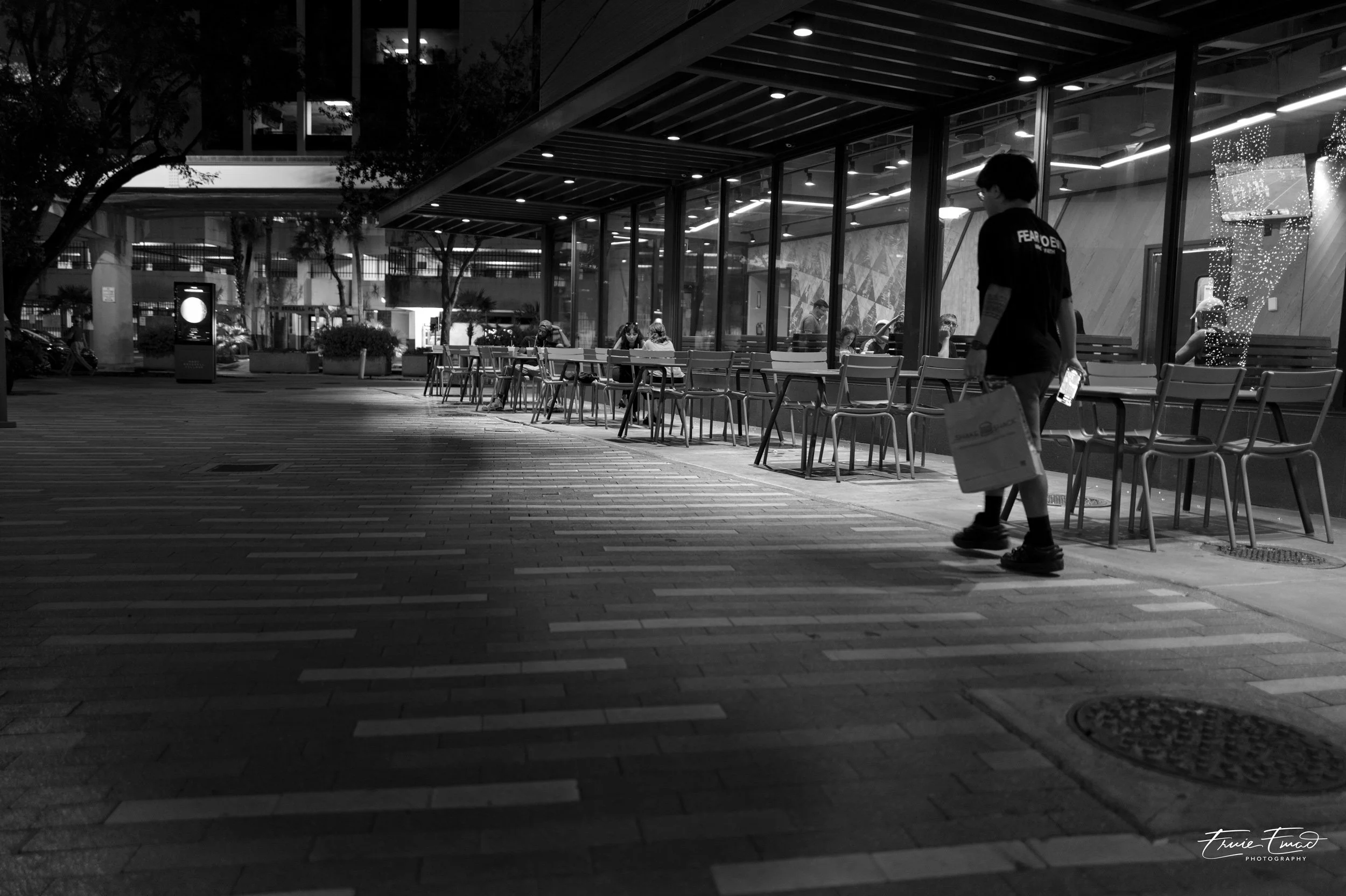 Black and white photo of a person walking past a modern building's outdoor seating area at night. The person is carrying a shopping bag and holding a phone, wearing a shirt that says 'Fear of' on the back. Inside the building, several people are sitt