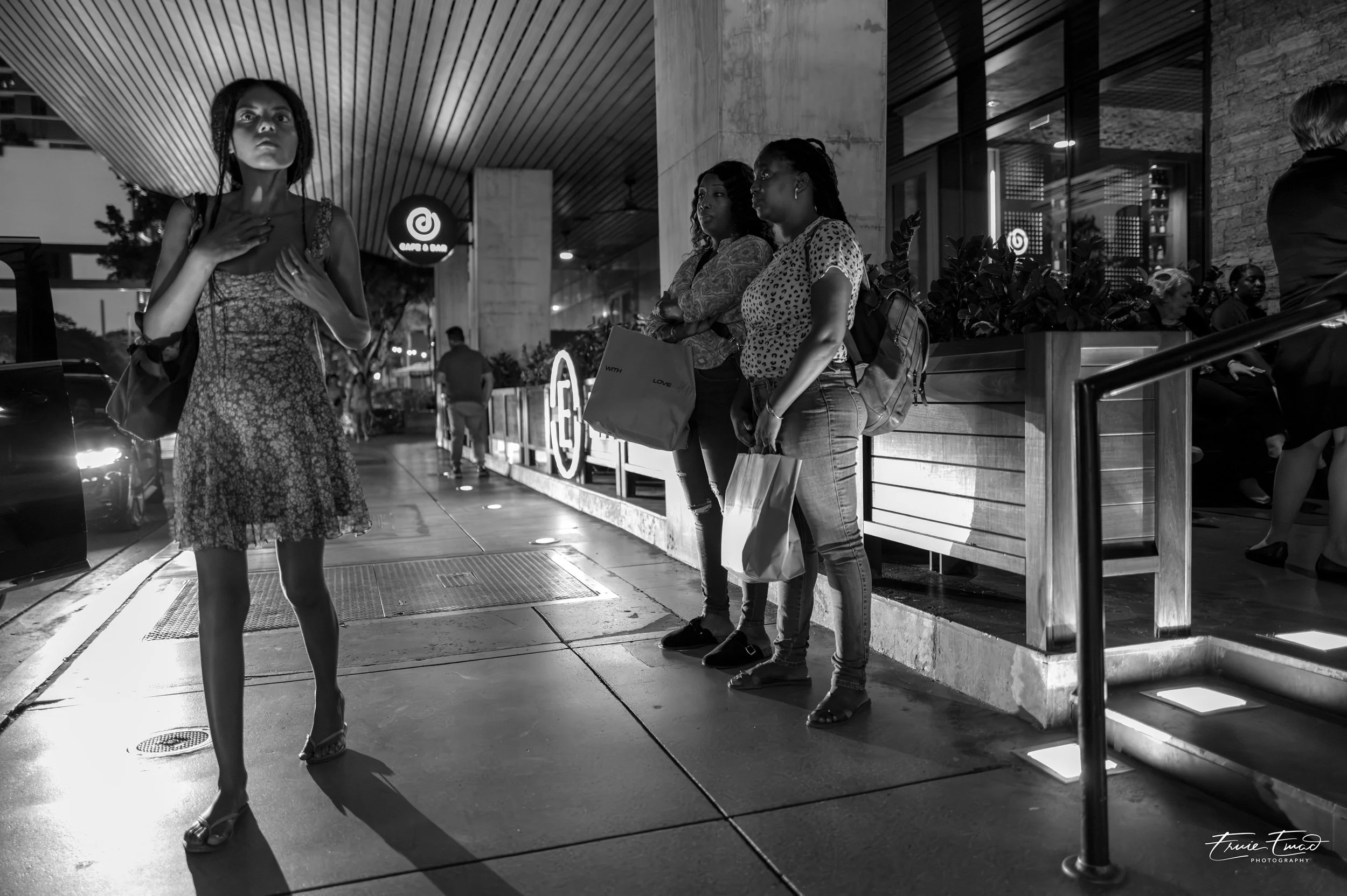 A group of women standing outside a restaurant in the evening. One woman in a floral dress is walking by, and three women are waiting in line, carrying shopping bags. The restaurant has a modern exterior with glass, brick, and wood elements, and a li