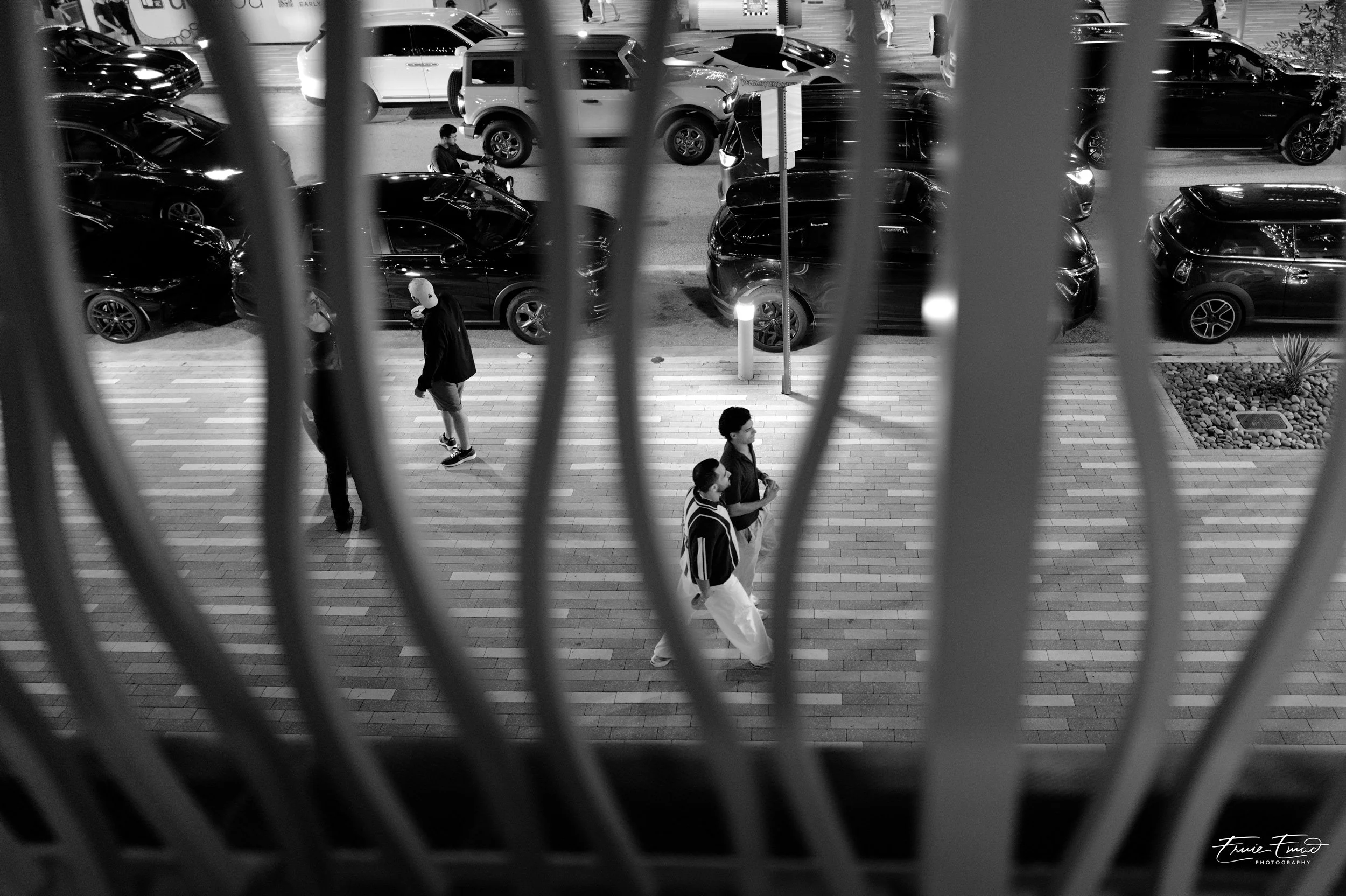 Black and white photo of a city street, viewed through a metal fence with vertical bars. Several pedestrians are walking on the sidewalk, and multiple cars are parked along the street. The scene appears to be at night or in low light conditions.