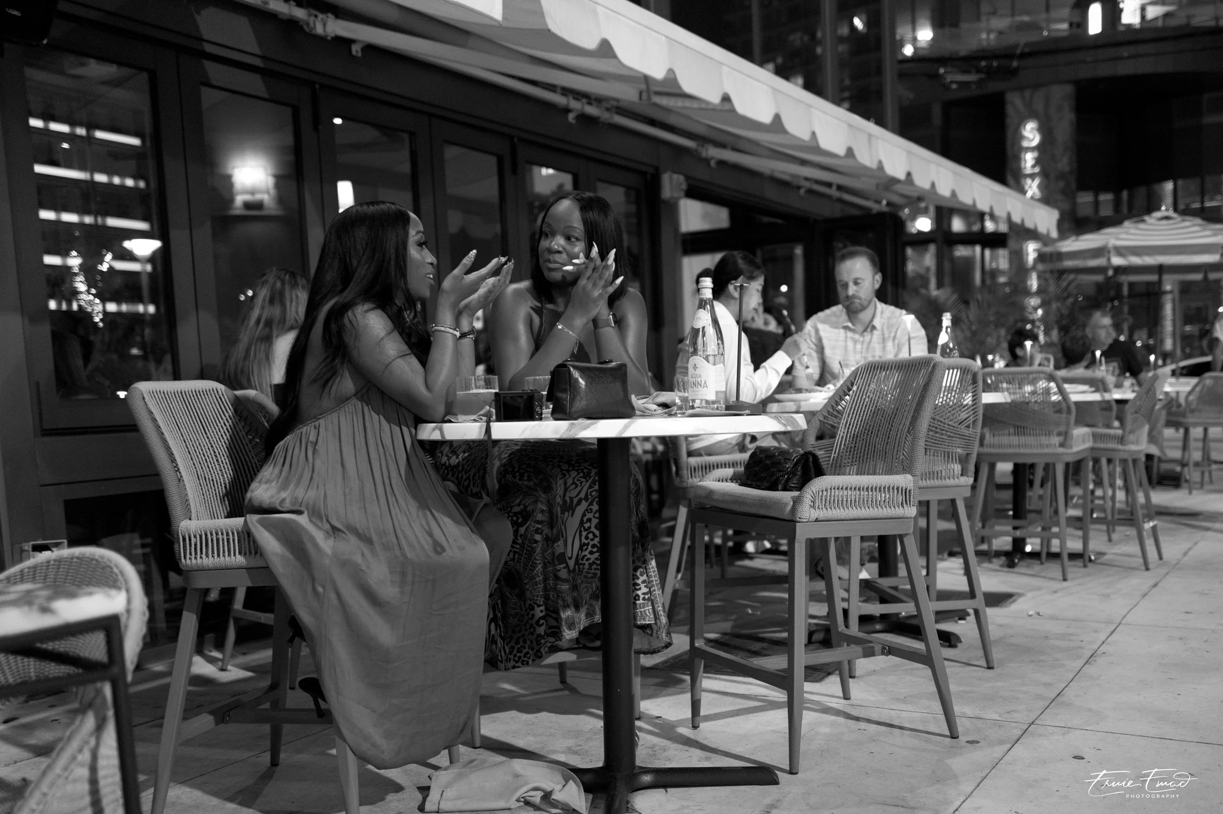 Two women sitting at an outdoor restaurant table, engaged in conversation, with bottles of water and glasses in front of them, nighttime setting.