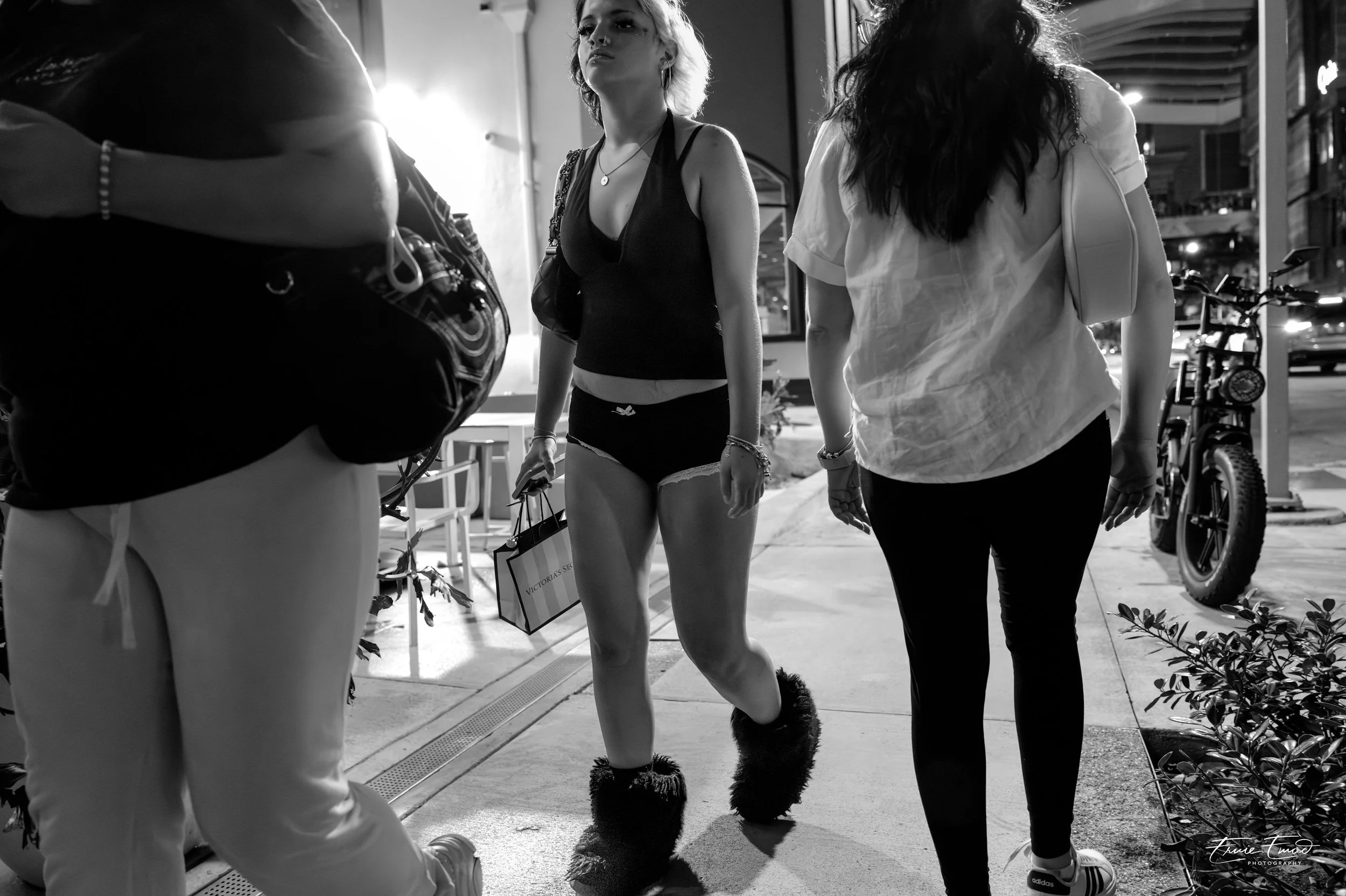 A black-and-white photo of three young women walking on a city sidewalk at night. One woman is wearing furry boots, a sleeveless top, and shorts, holding a shopping bag. Another woman is dressed in a white shirt with rolled-up sleeves, and the third 