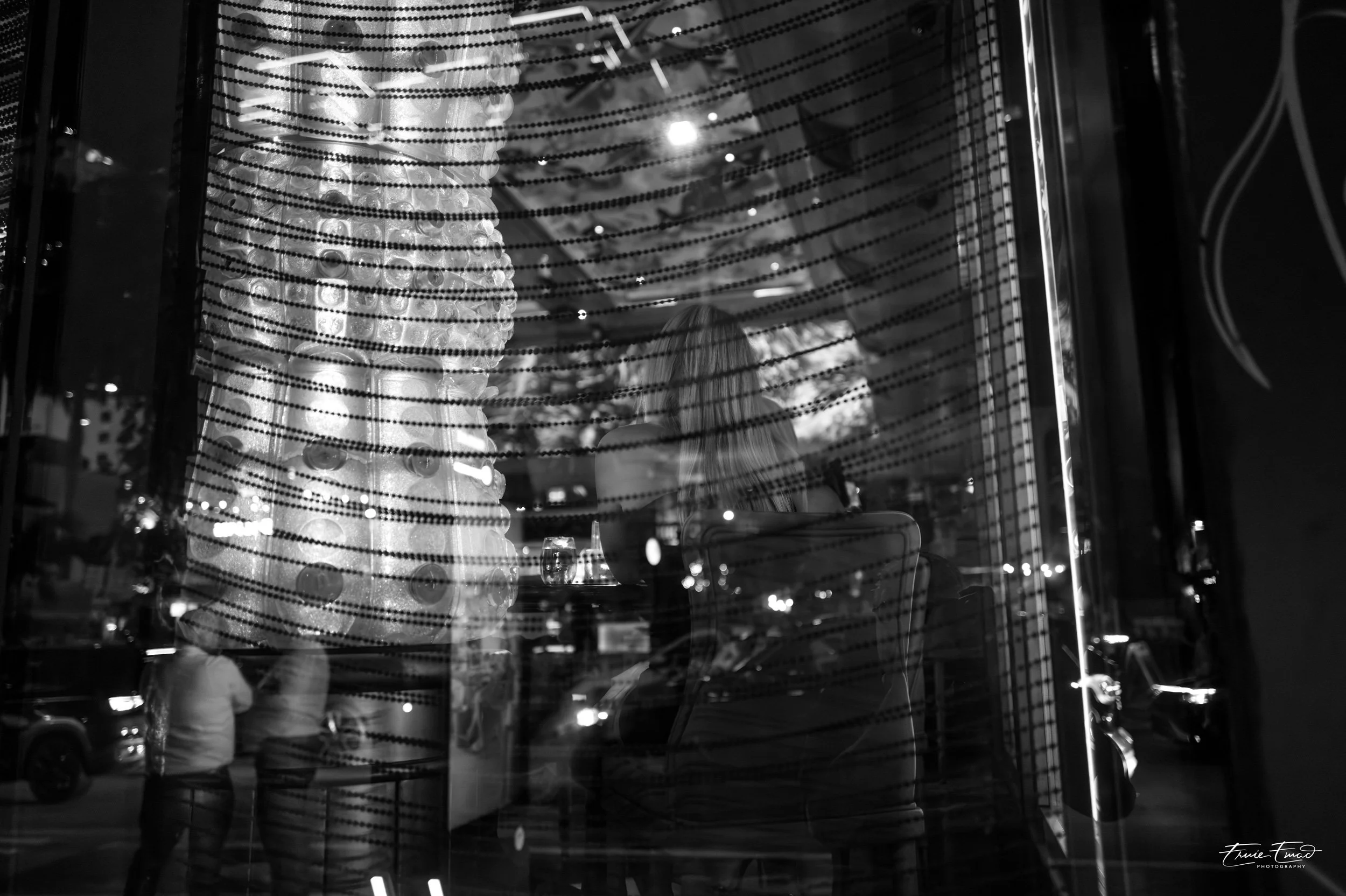 People sitting at tables inside a restaurant or café, seen through a window with horizontal blinds, at night.