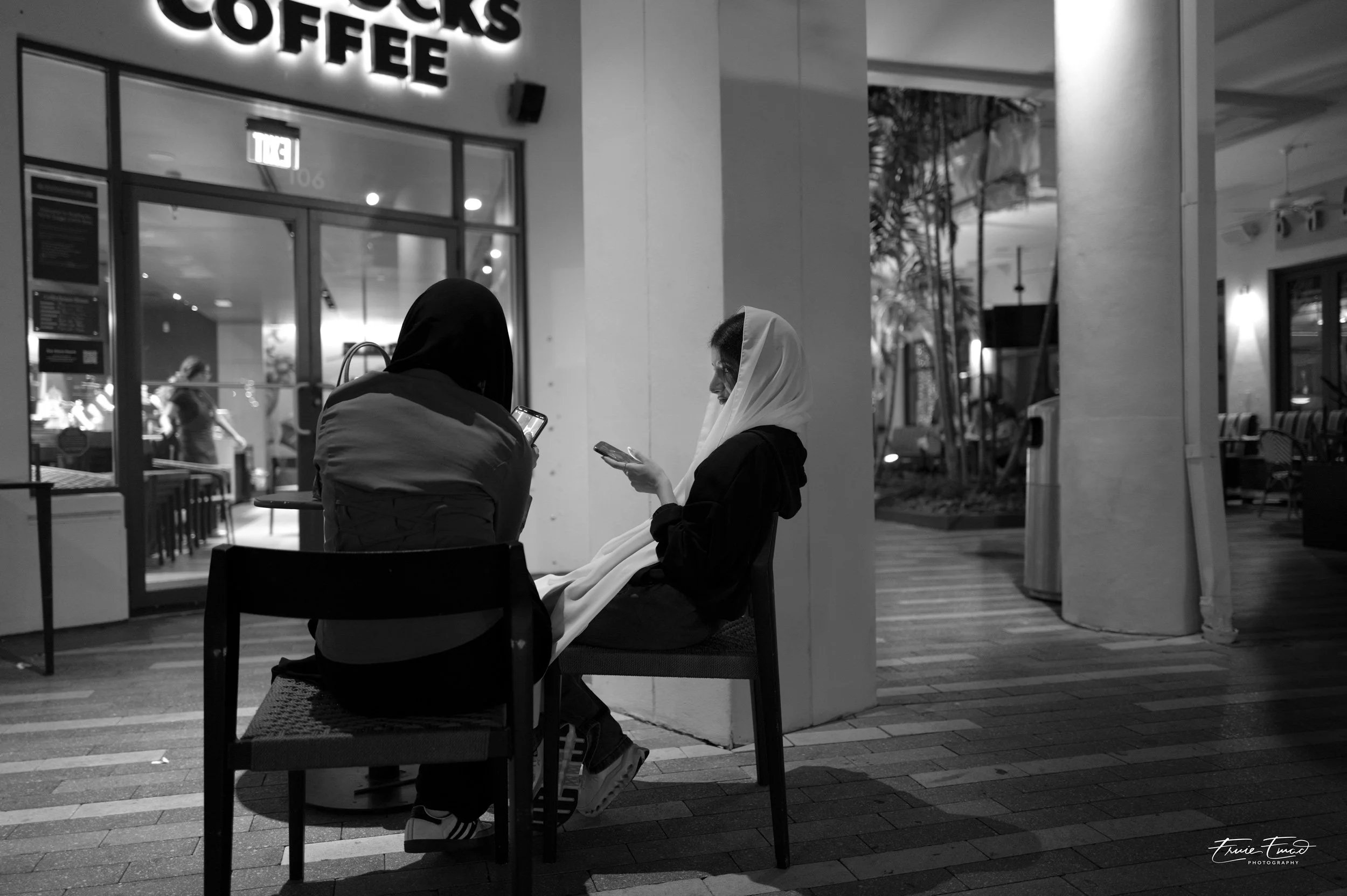 Two women sitting on chairs outside a coffee shop at night, one wearing a dark hijab and the other a light hijab, both looking at their phones. The coffee shop has illuminated signs and glass doors, with indoor lighting and seating visible through th