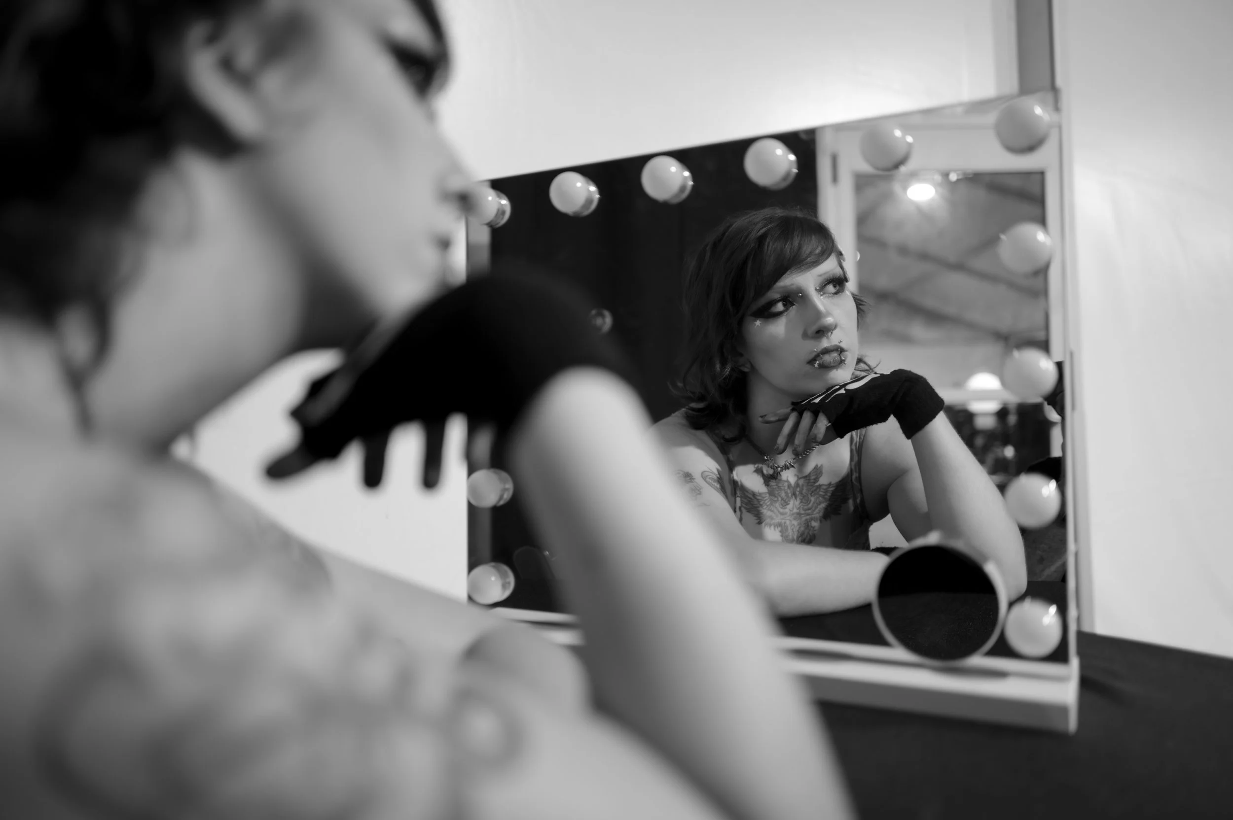 A person with short hair and makeup looking into a mirror with light bulbs around it, reflecting their face and arm with tattoos. At Sounds of Screams Miami