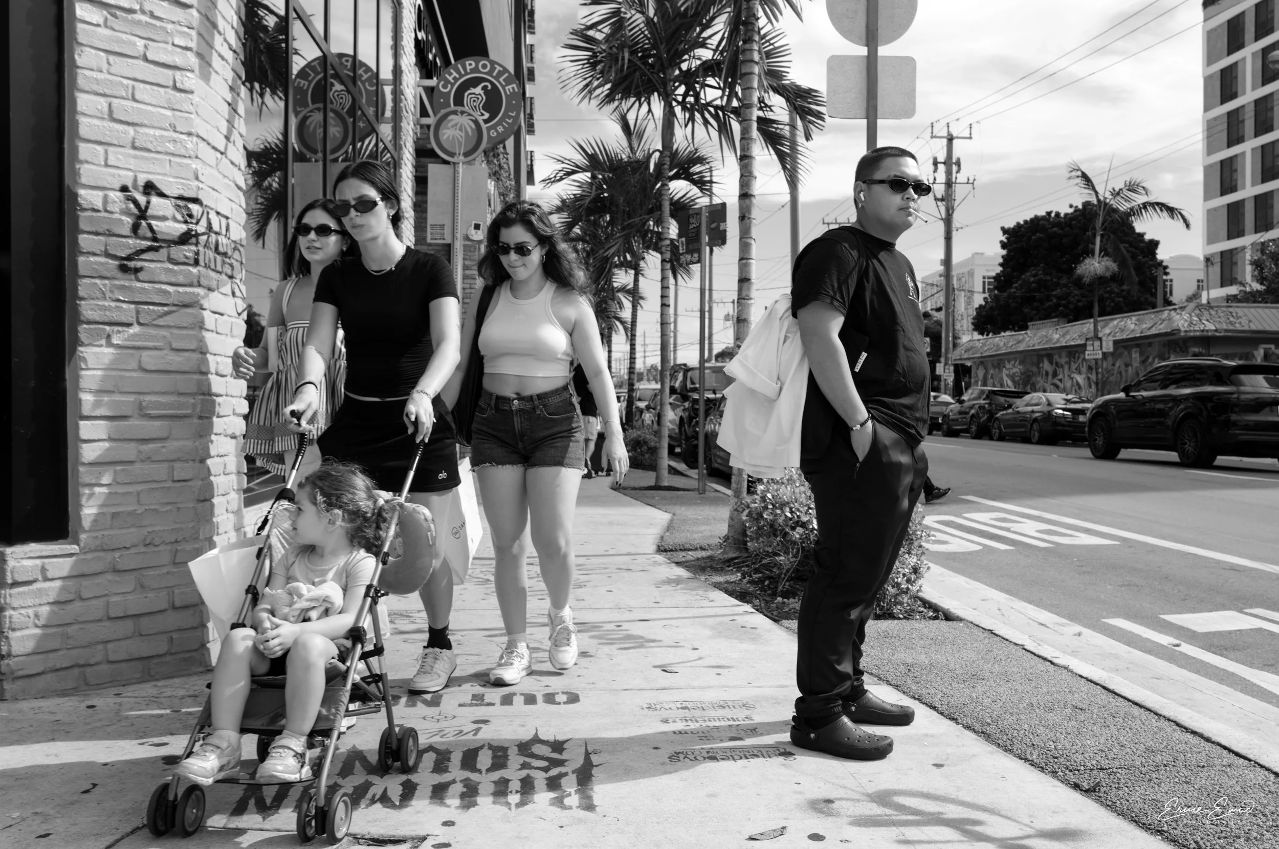 A group of five people walking along a sidewalk on a city street, with palm trees and parked cars visible in the background. One woman is pushing a stroller with a young girl sitting inside. The others are walking alongside, all wearing sunglasses. A