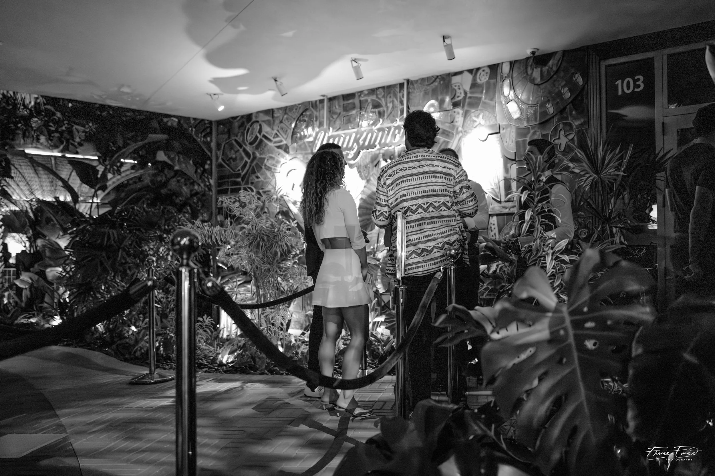 Black and white photo of four people seen from behind standing in line inside a venue with plants, a decorative mural, and a neon sign that says 'Chill Zone'. The atmosphere appears indoor and social.